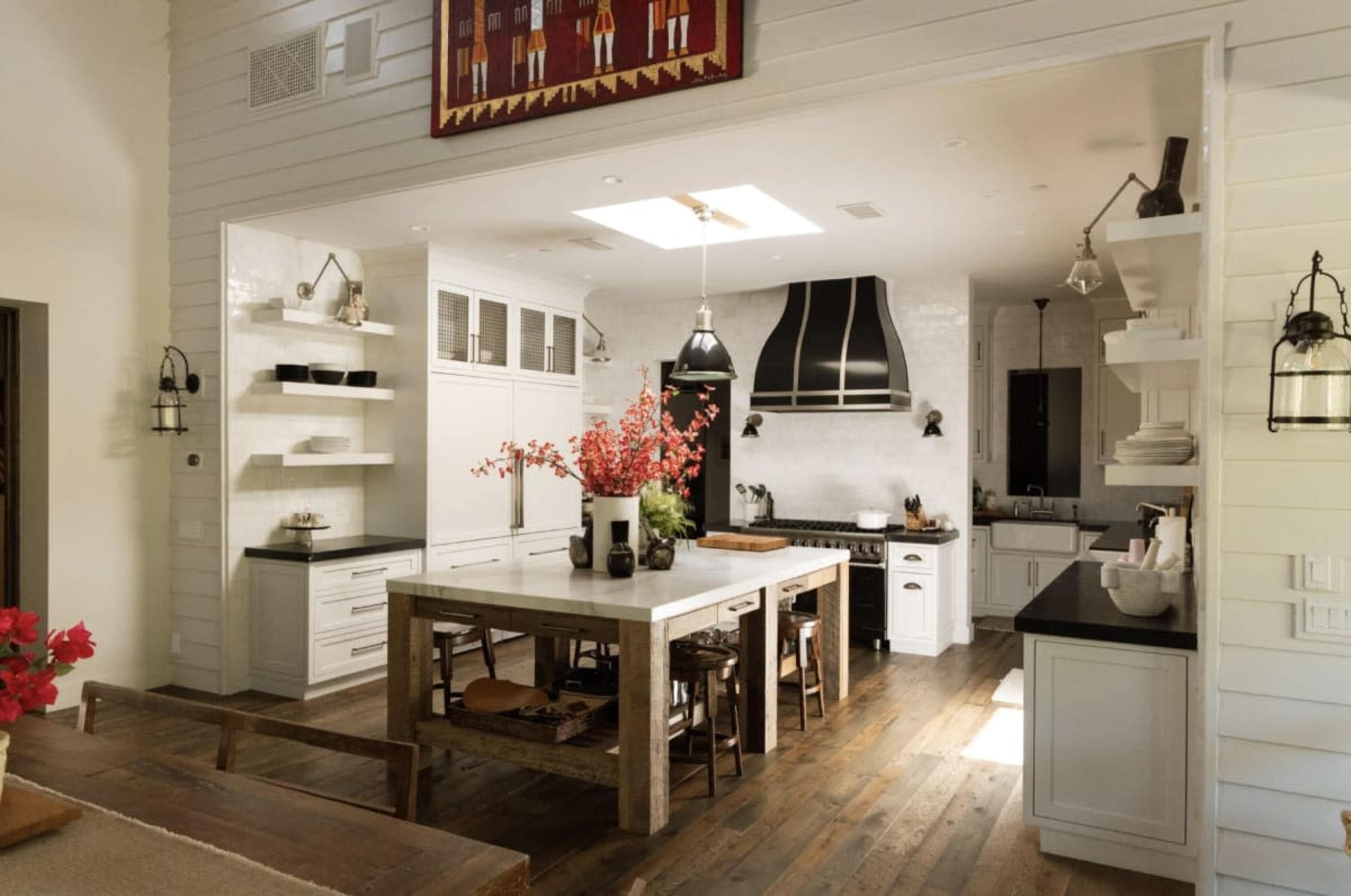 The image shows a spacious kitchen with a central dining table, modern appliances, and white cabinetry, featuring shelves and decorative elements.
