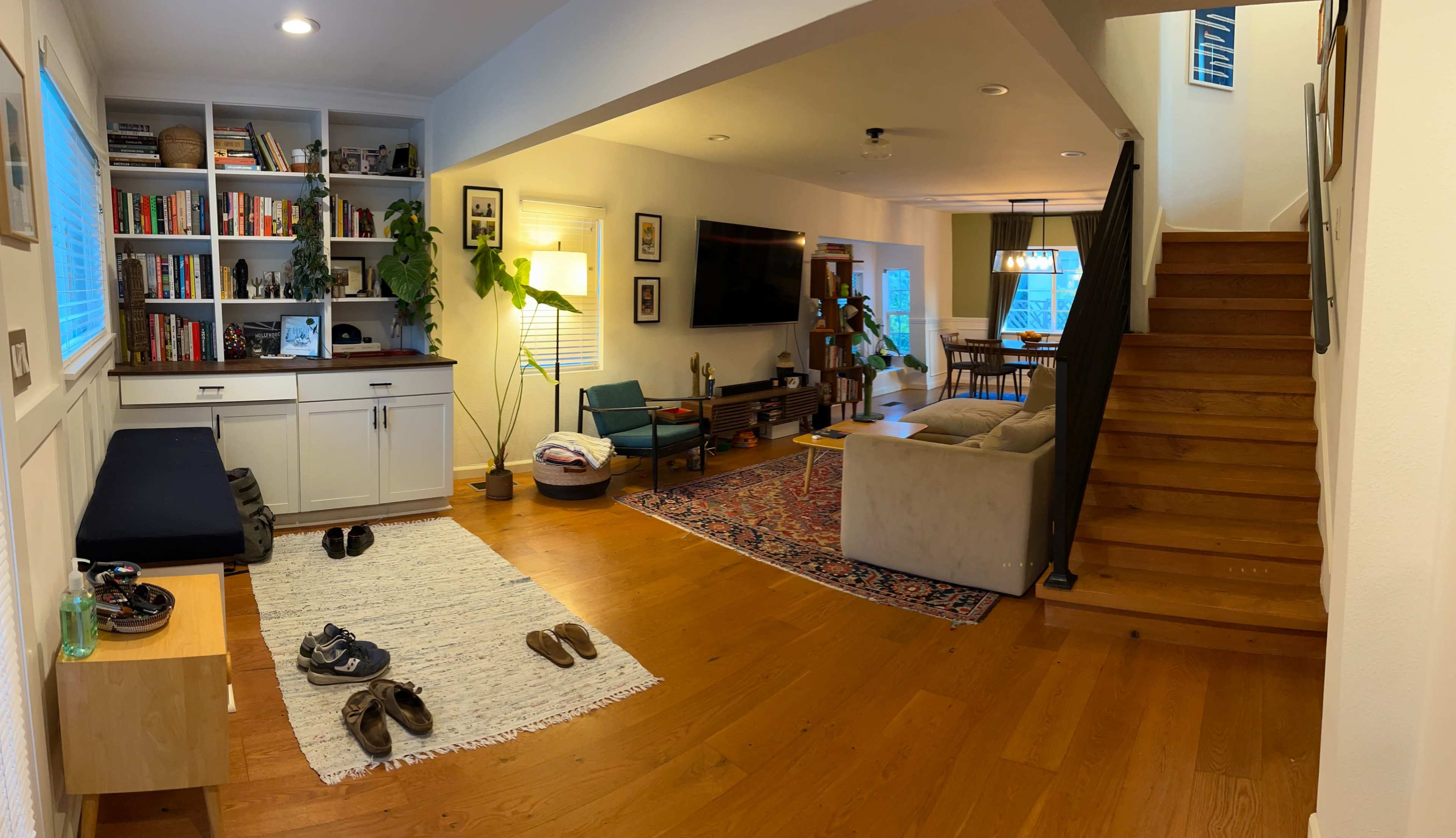 A spacious living area with wooden floors, a staircase, bookshelves, a television, a couch, and various indoor plants.
