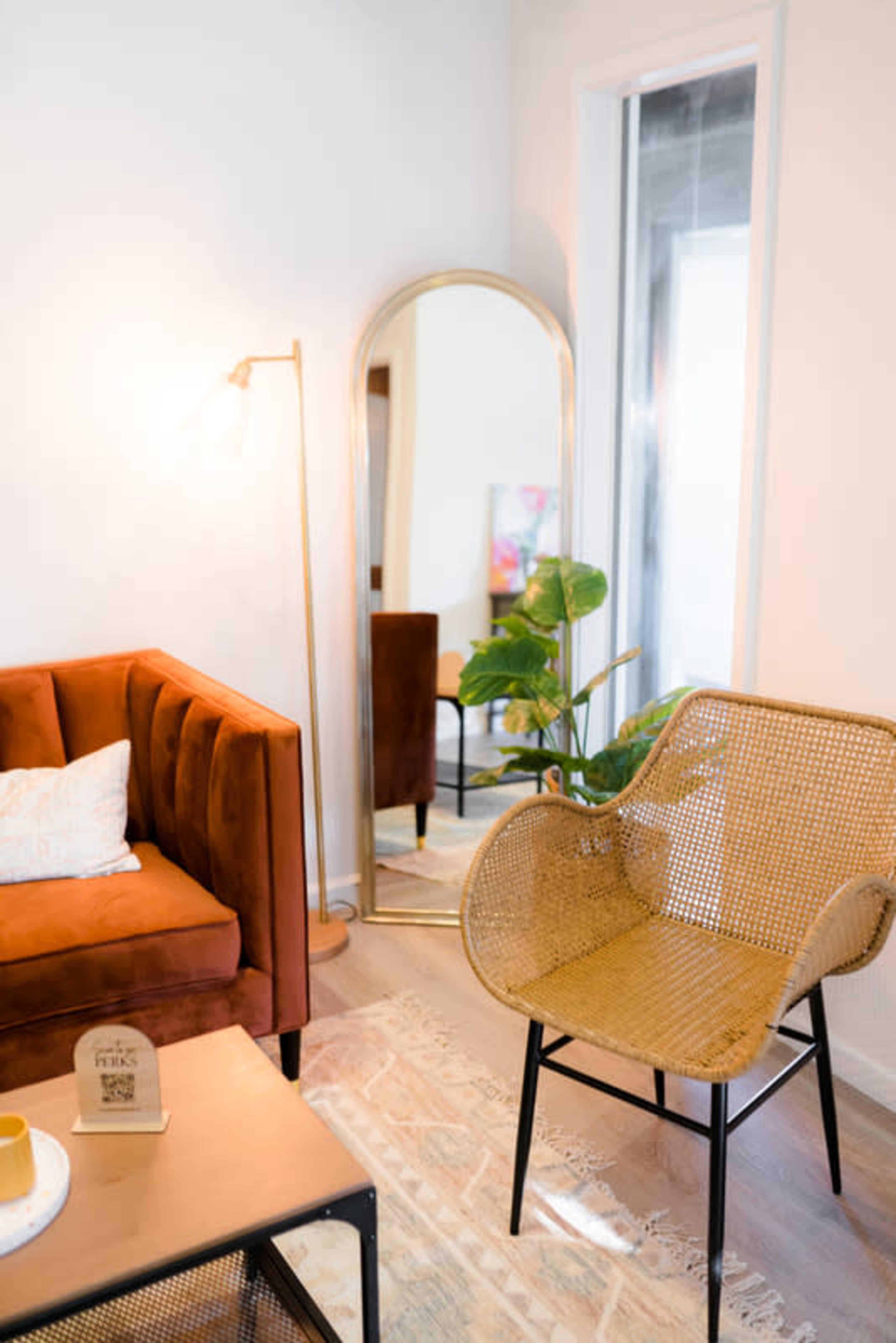 A cozy living room featuring a rust-colored sofa, a rattan chair, a gold-framed mirror, and a green plant beside a coffee table.