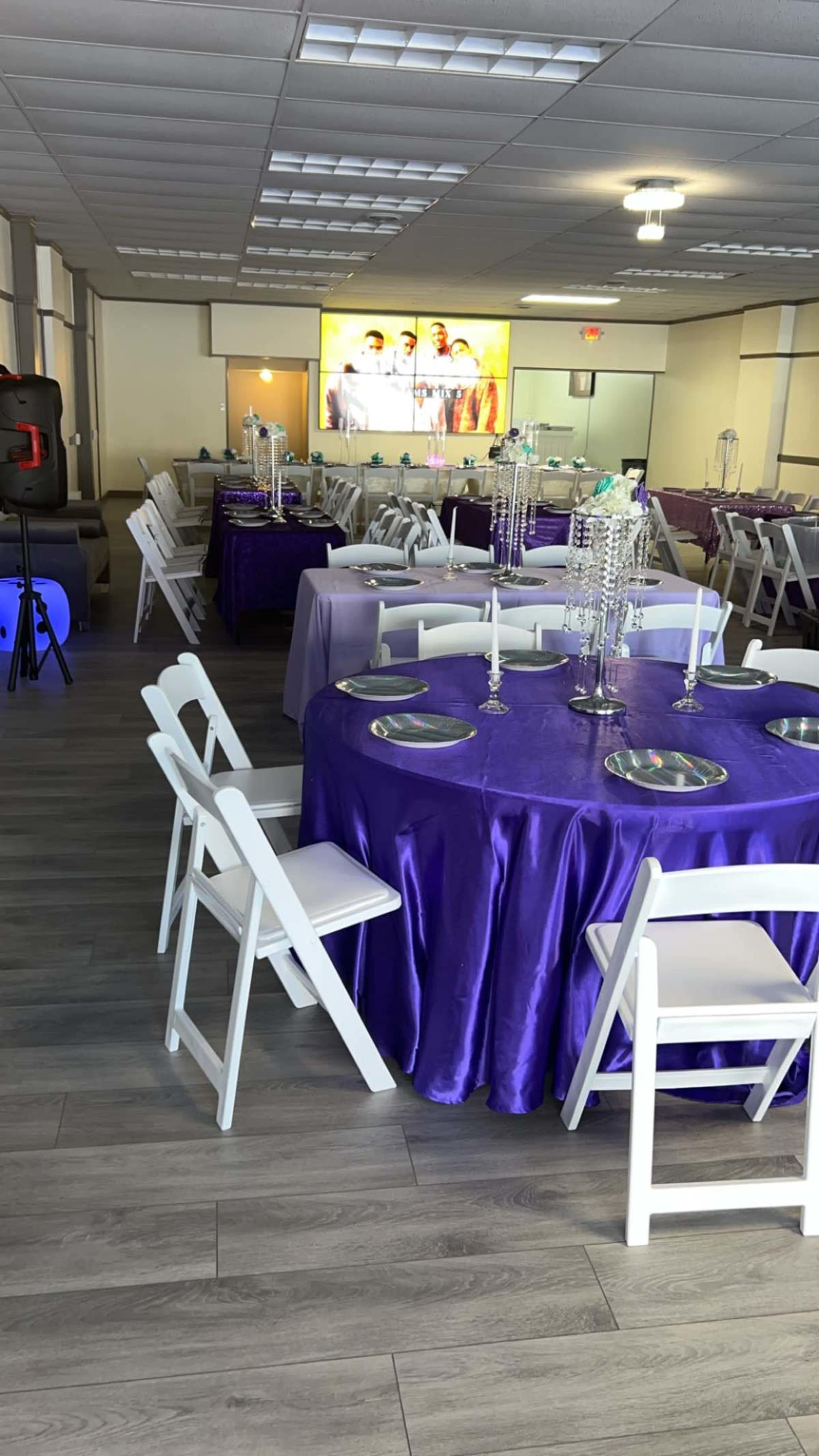 The image shows a banquet-style room set up with several tables covered in purple tablecloths, surrounded by white chairs, and a screen displaying an image in the background.