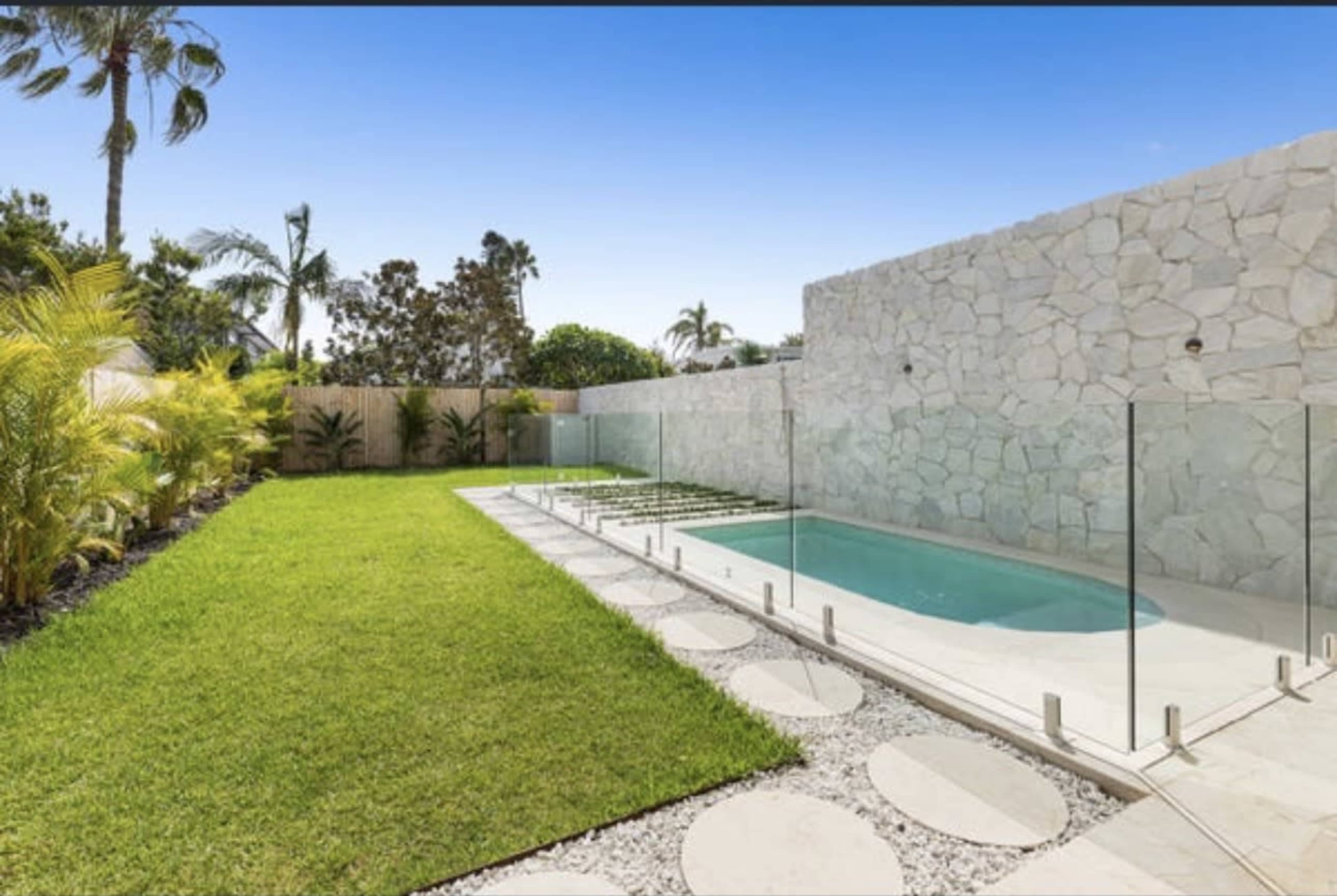 The image shows a landscaped backyard with a small swimming pool surrounded by glass fencing, neatly trimmed grass, and stone pathways.