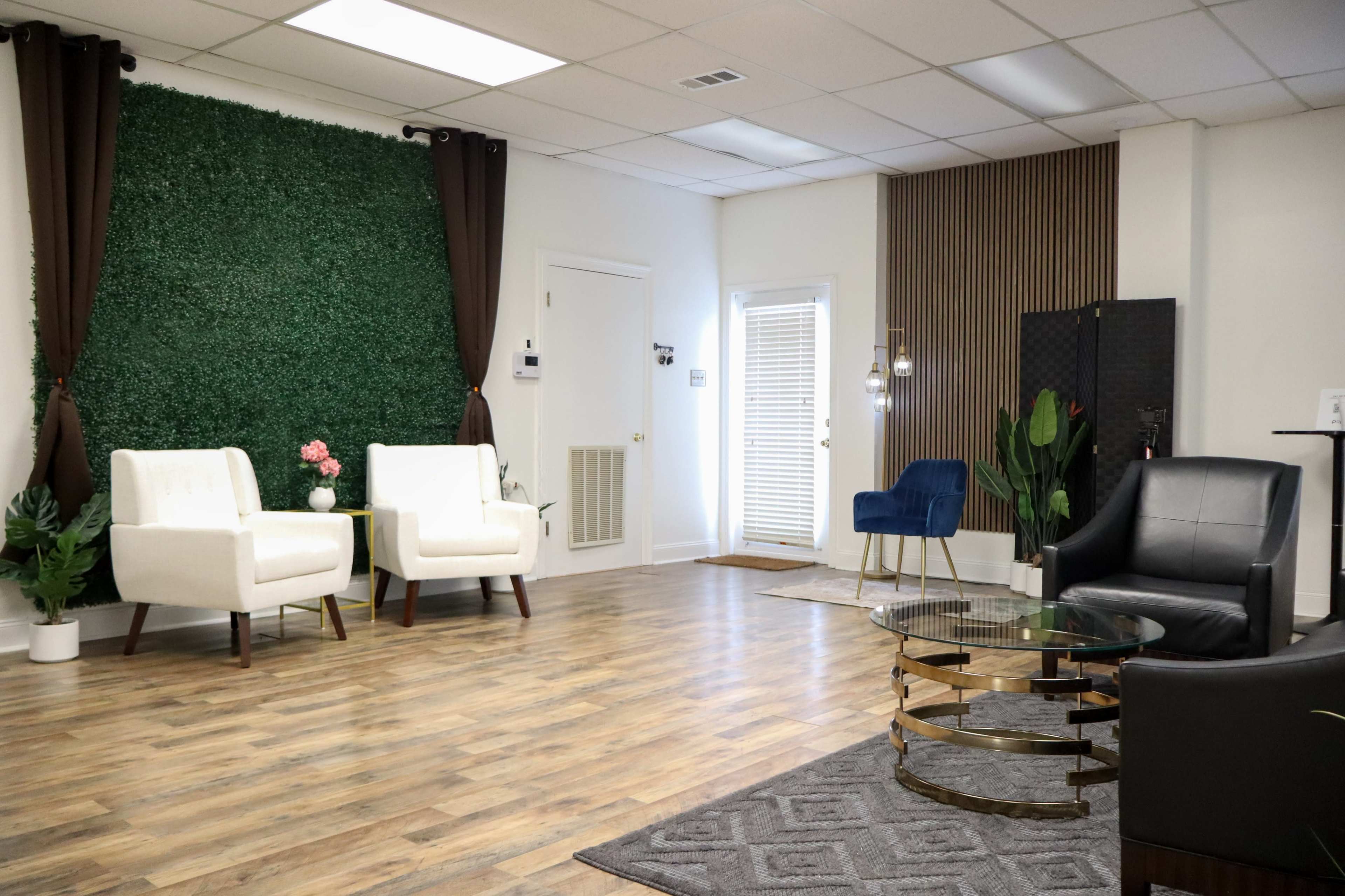 The image shows a modern waiting area with two white chairs, a blue chair, a round coffee table, and decorative plants against a backdrop of green foliage.