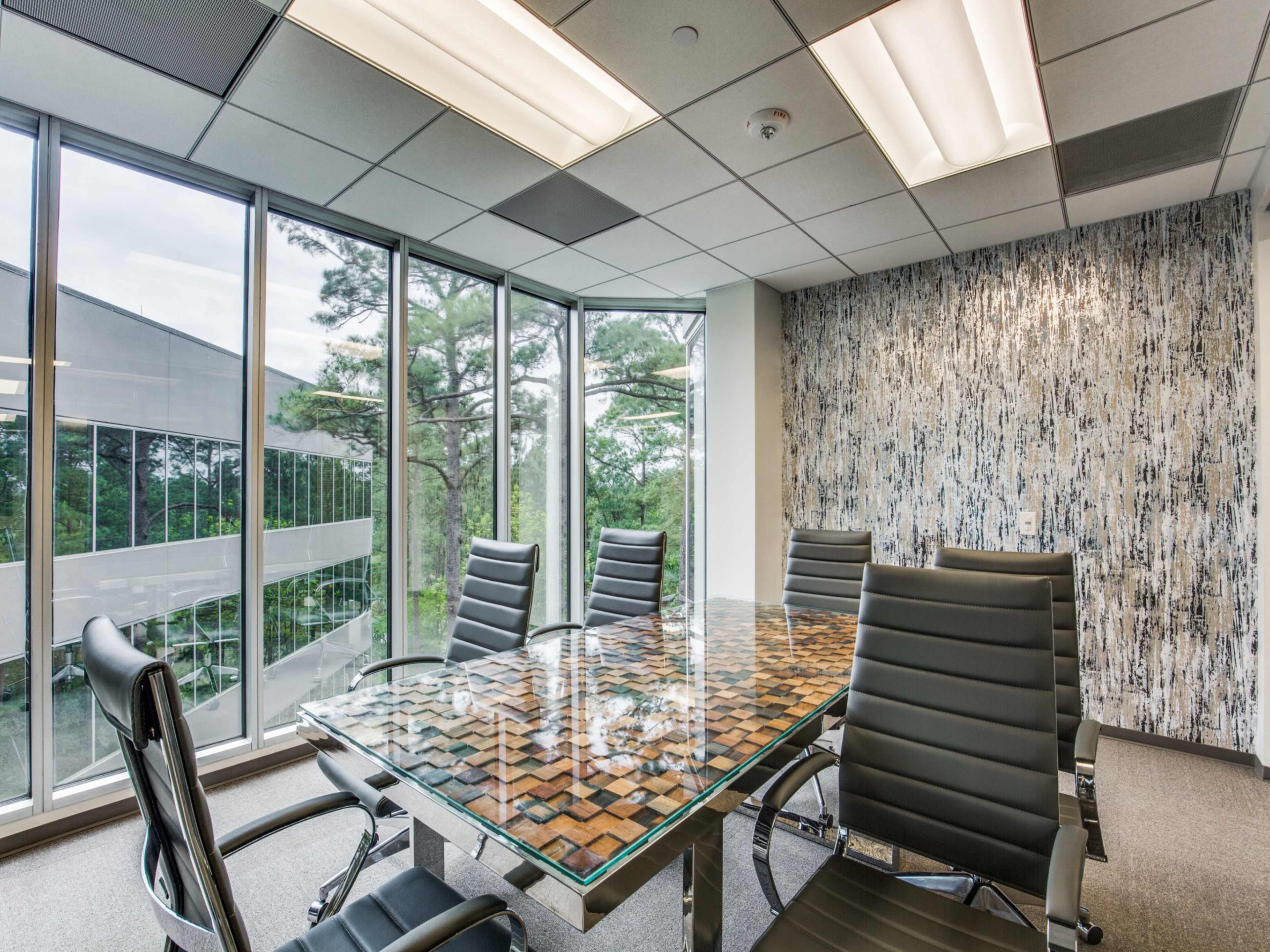 A conference room features a glass-topped table surrounded by black leather chairs, with large windows displaying a view of trees.