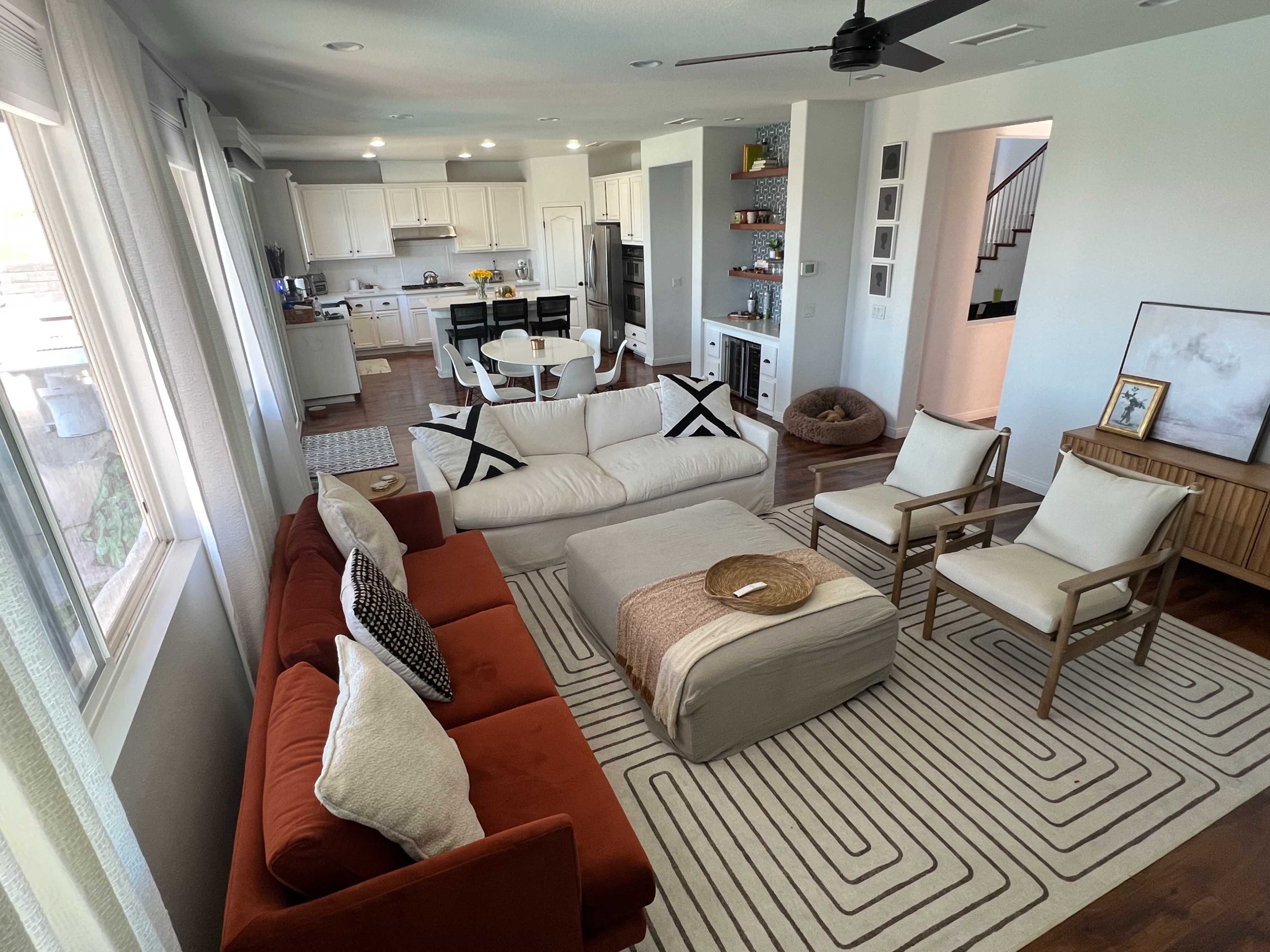 The image shows a modern living room with an orange sectional sofa, a central coffee table, and light-colored armchairs arranged around a patterned area rug, with an open kitchen visible in the background.