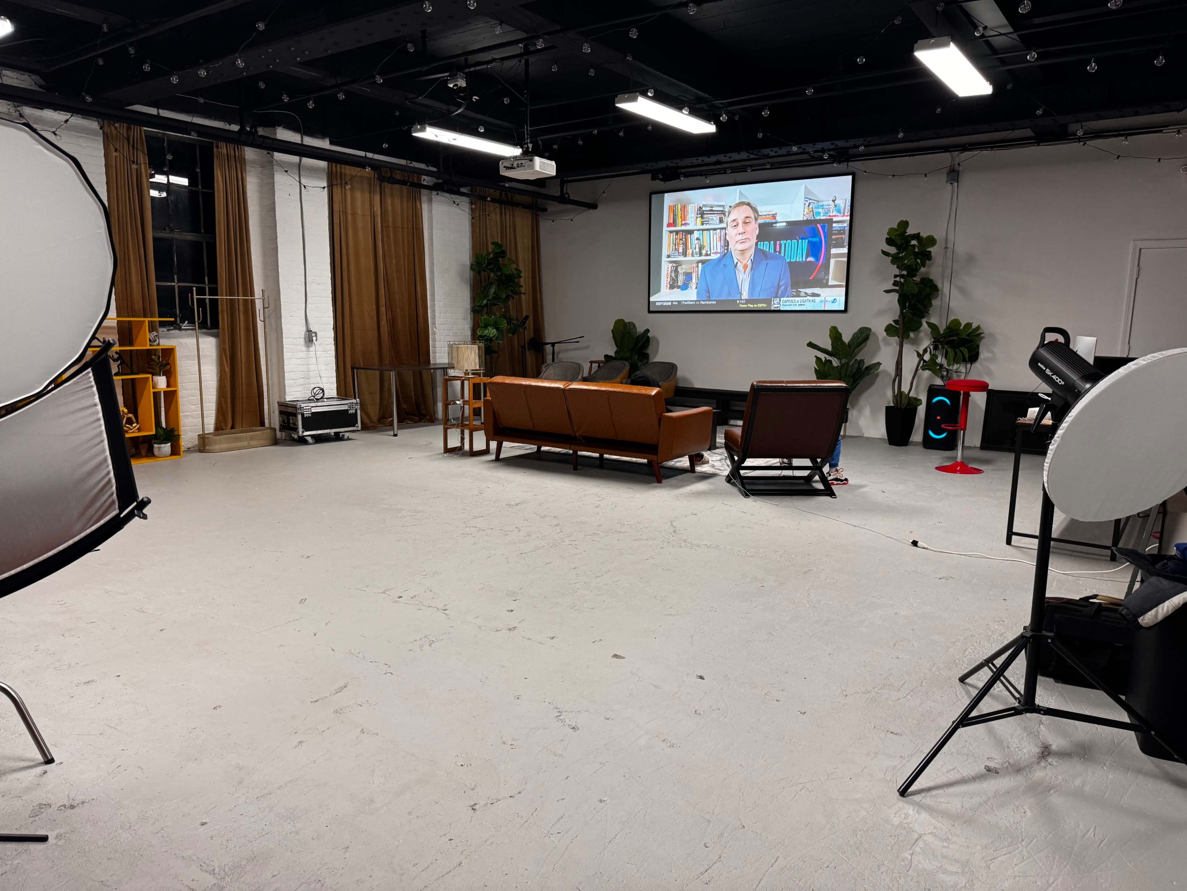 The image shows a spacious studio with two brown leather sofas, a large screen displaying a man, and various lighting equipment scattered around.