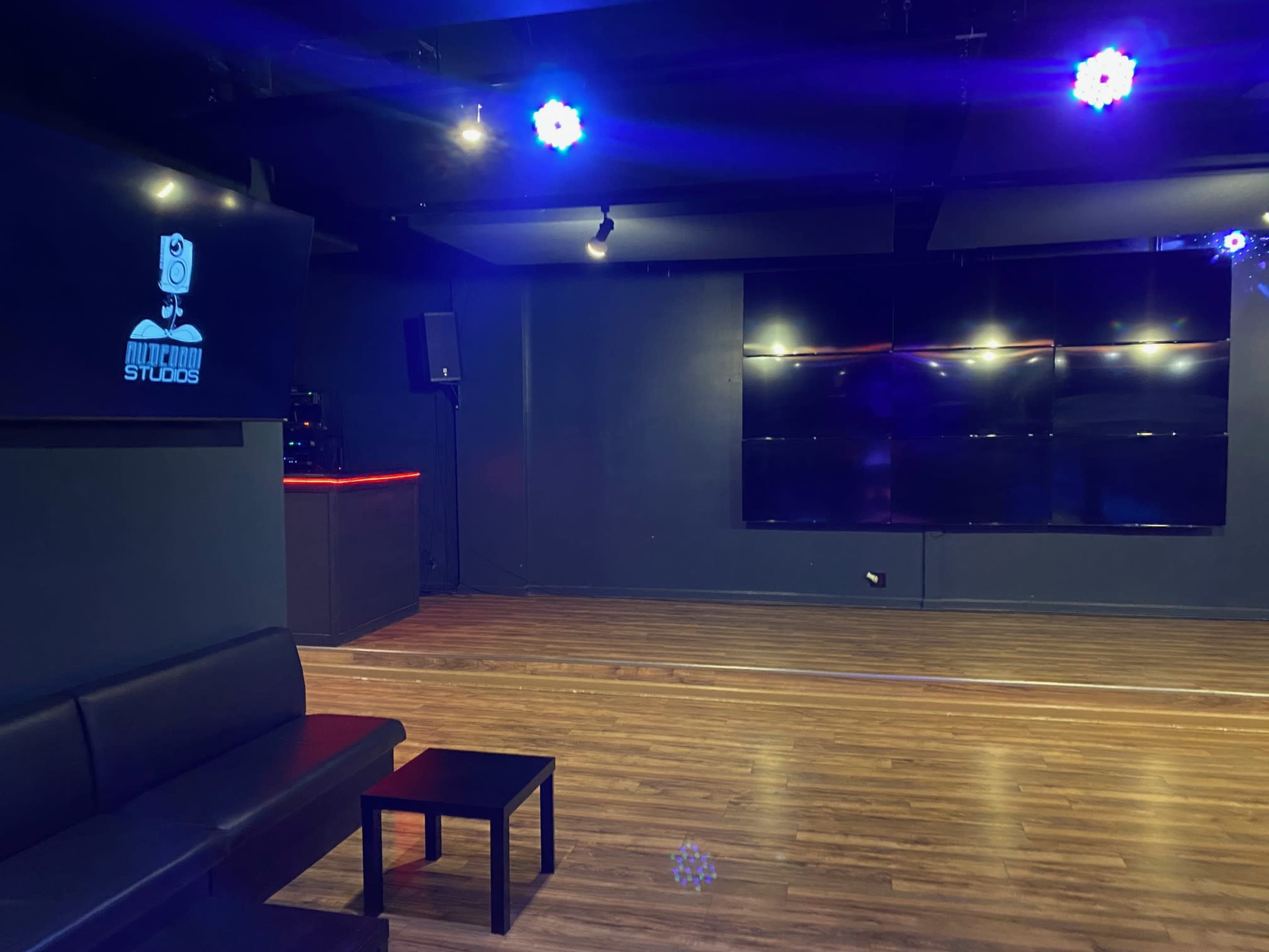 A music studio with a black couch and coffee table, illuminated by blue lights and featuring a large screen on the wall.