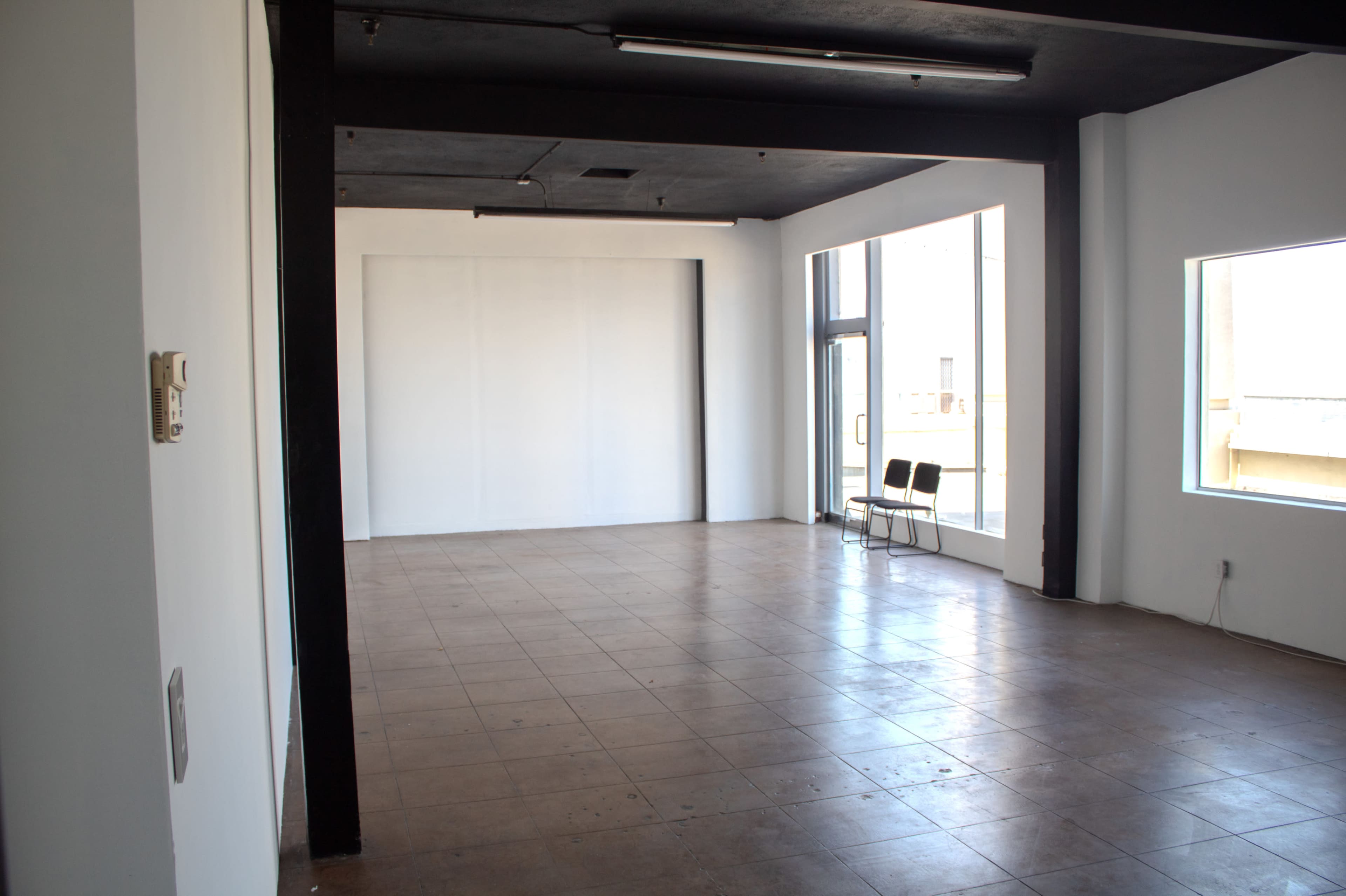 The image shows an empty room with tiled flooring, white walls, and large windows, featuring two chairs near the entrance.
