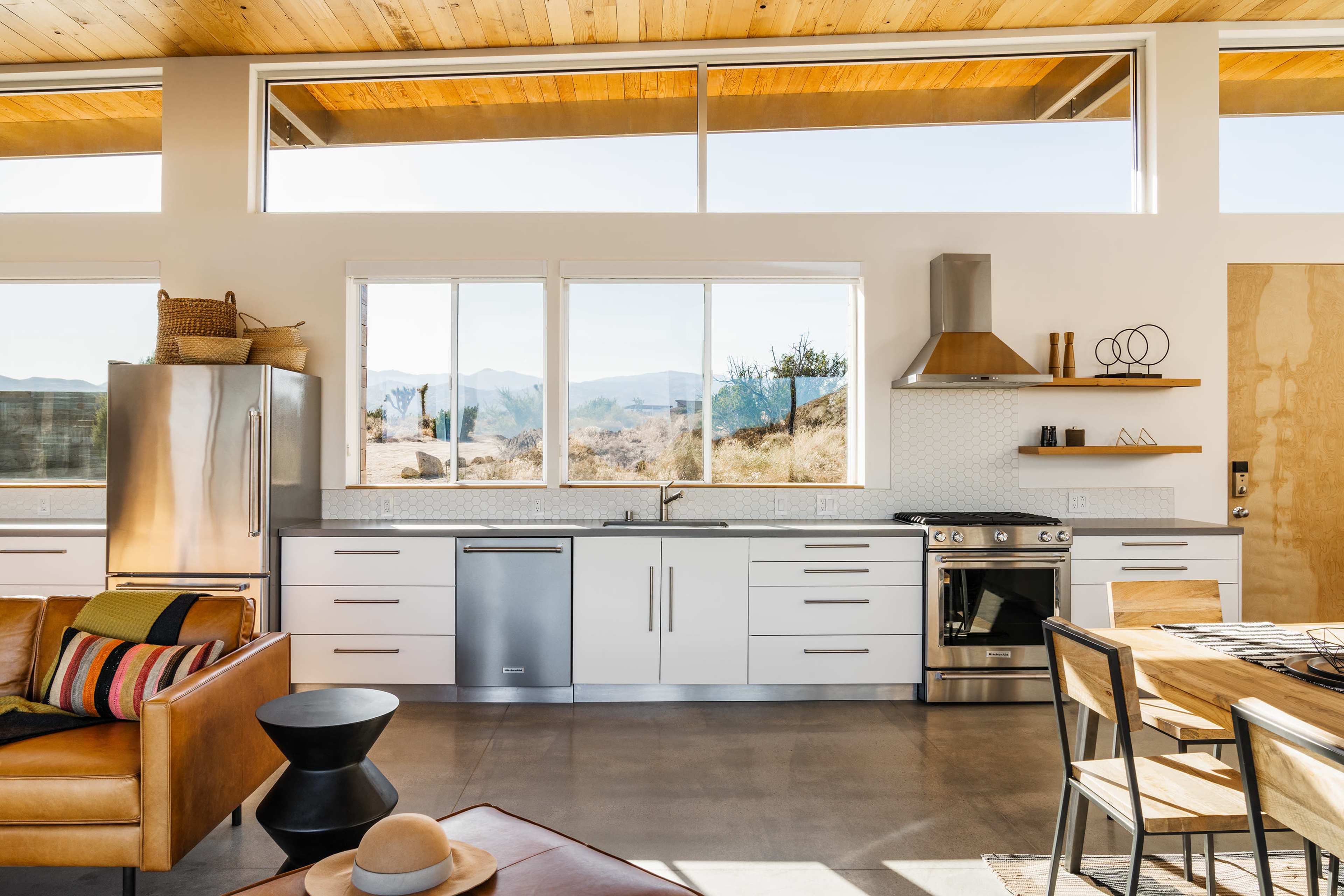 The image shows a modern kitchen with a stainless steel refrigerator, a gas stove, and large windows providing a view of a desert landscape.