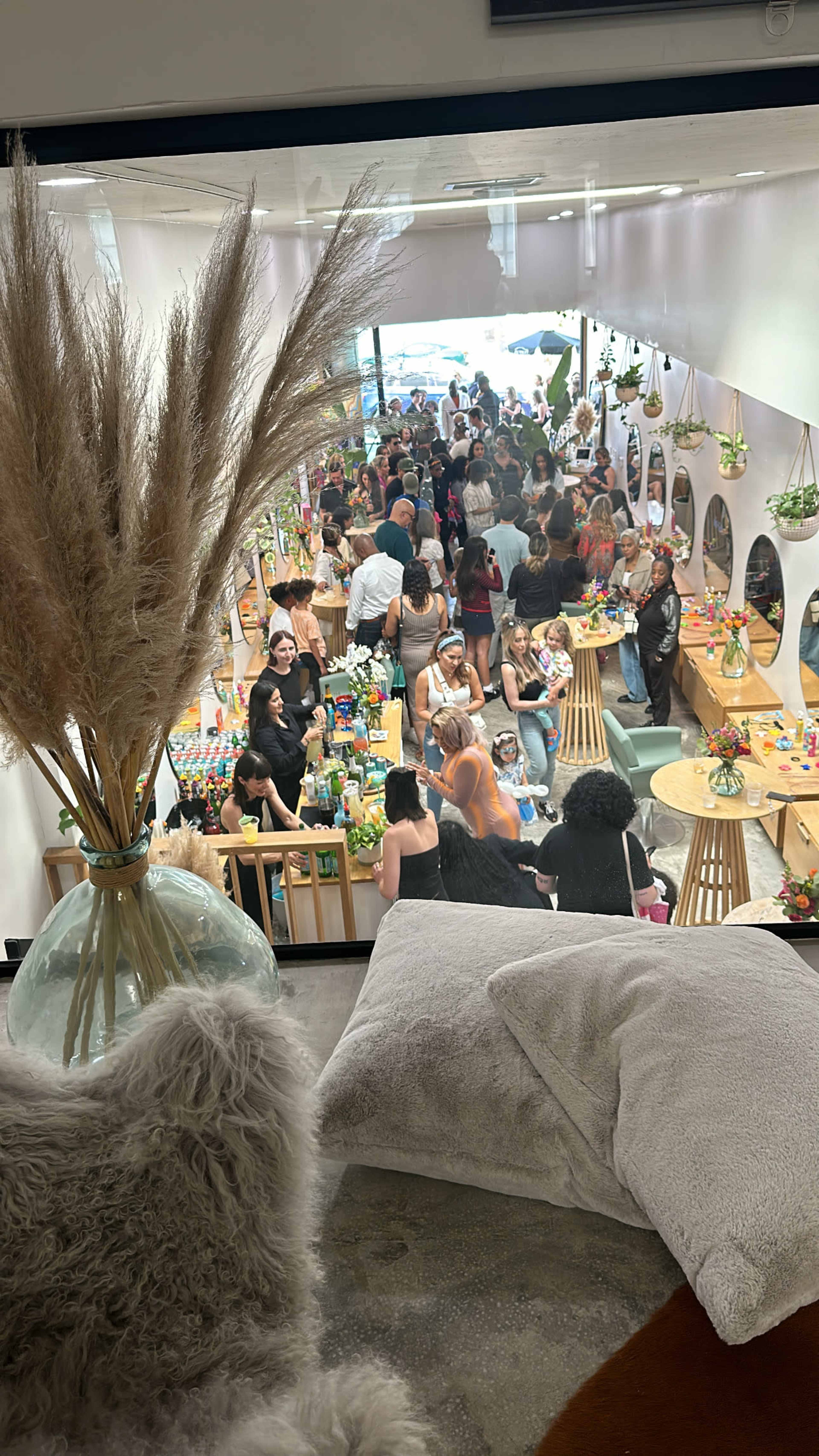 A bustling indoor event features a large crowd socializing around tables adorned with colorful drinks and decorations.