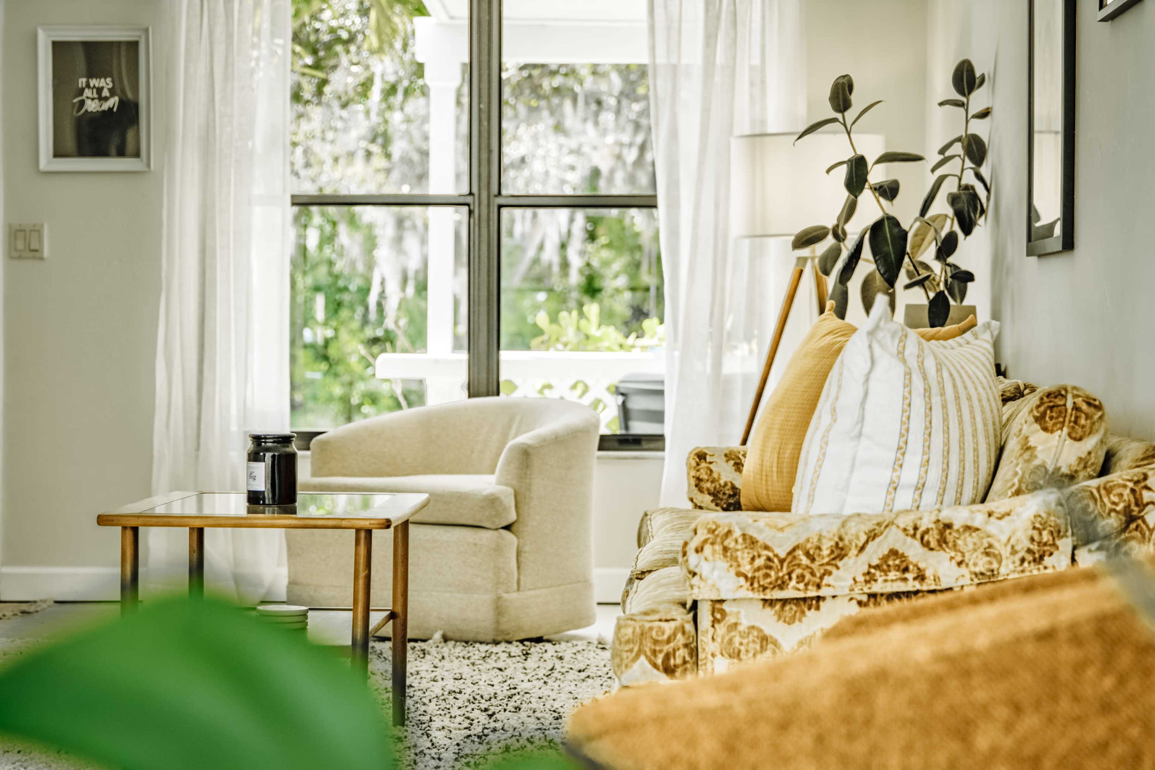 A beige sofa and armchair are positioned in a brightly lit living room with large windows and green plants visible outside.