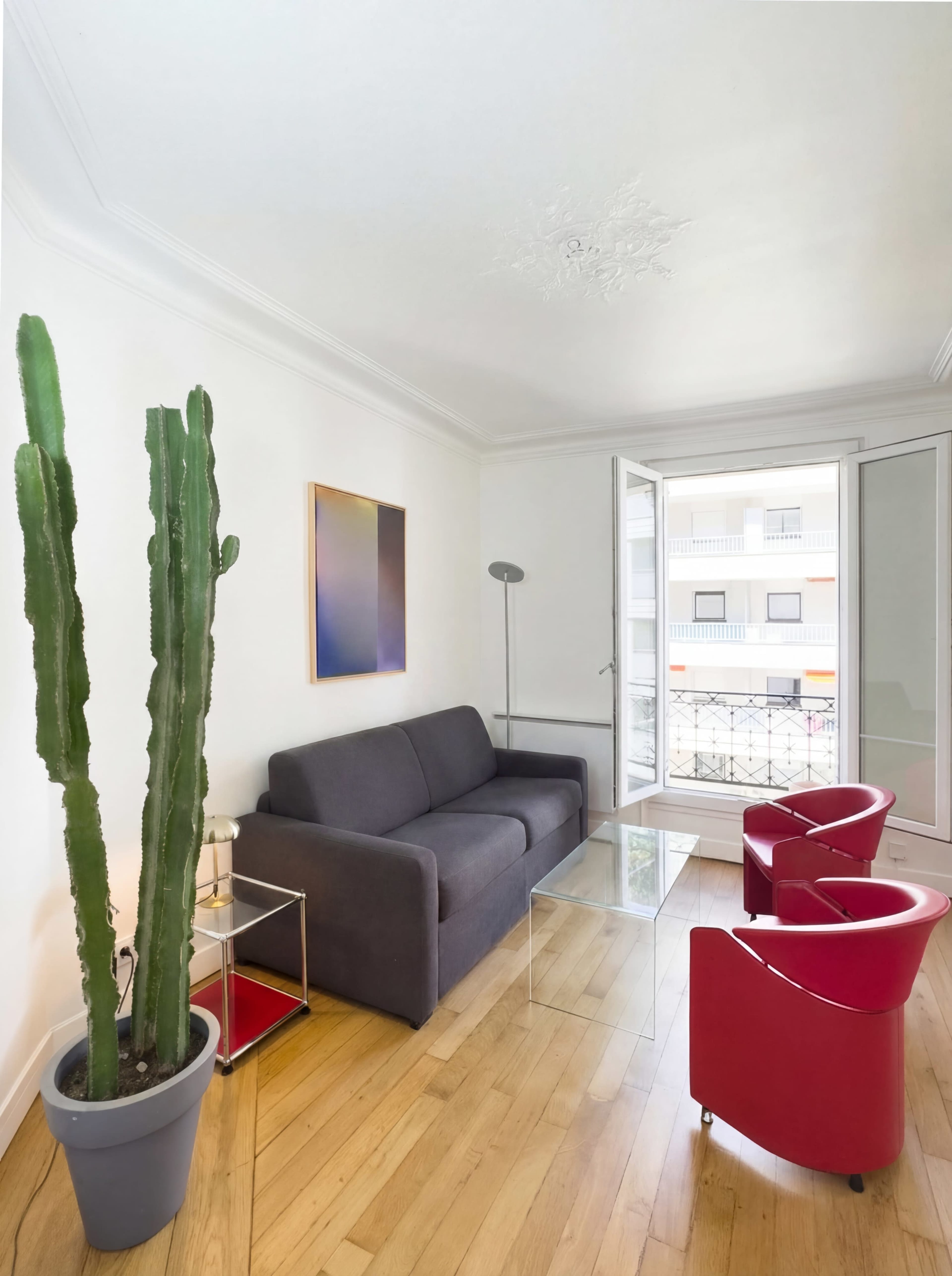 The image shows a modern living room with a gray couch, two red chairs, a glass coffee table, and a tall cactus in a planter next to an open balcony door.