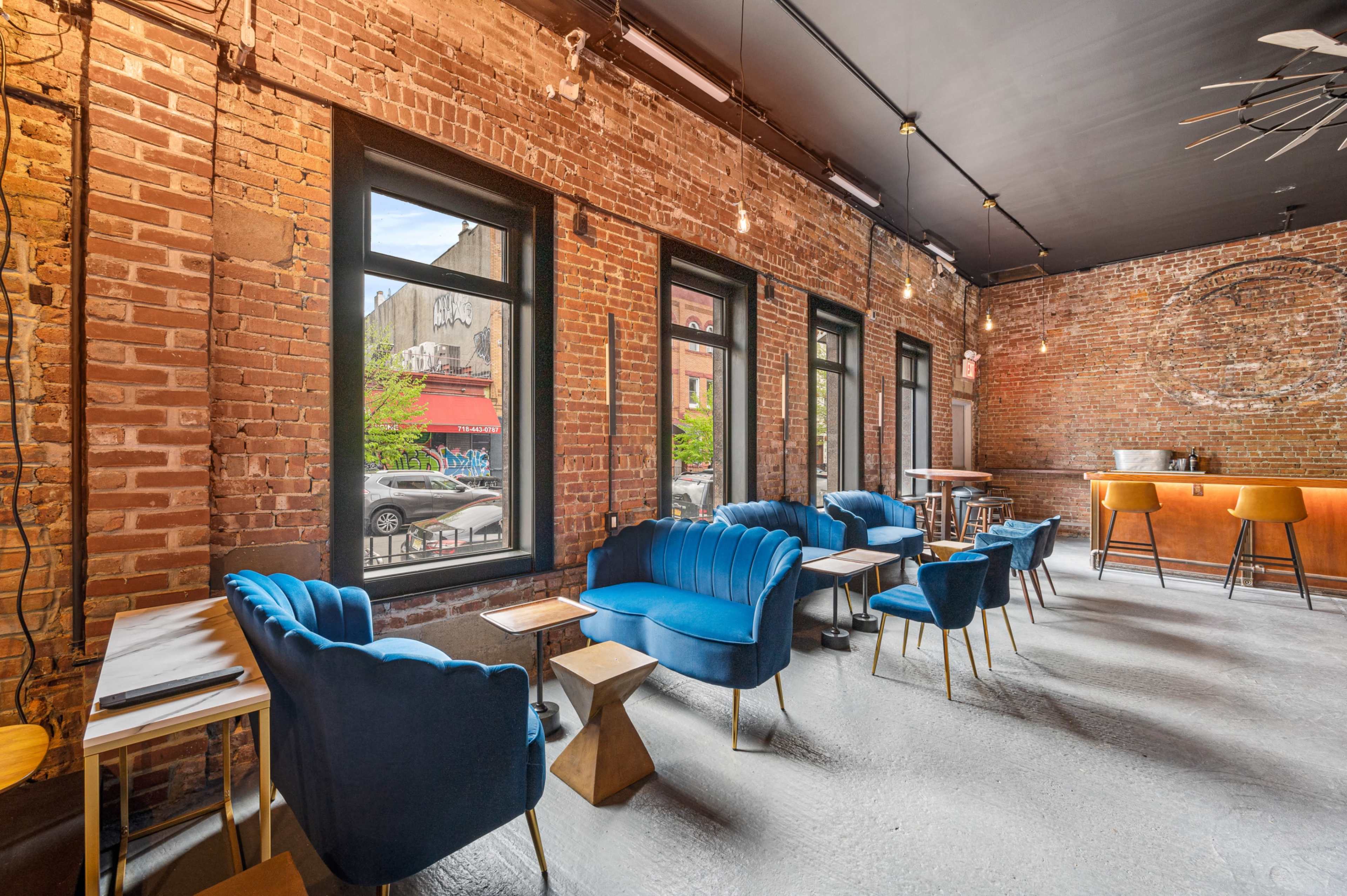 A café with exposed brick walls, blue upholstered chairs, small tables, and large windows looking out onto the street.