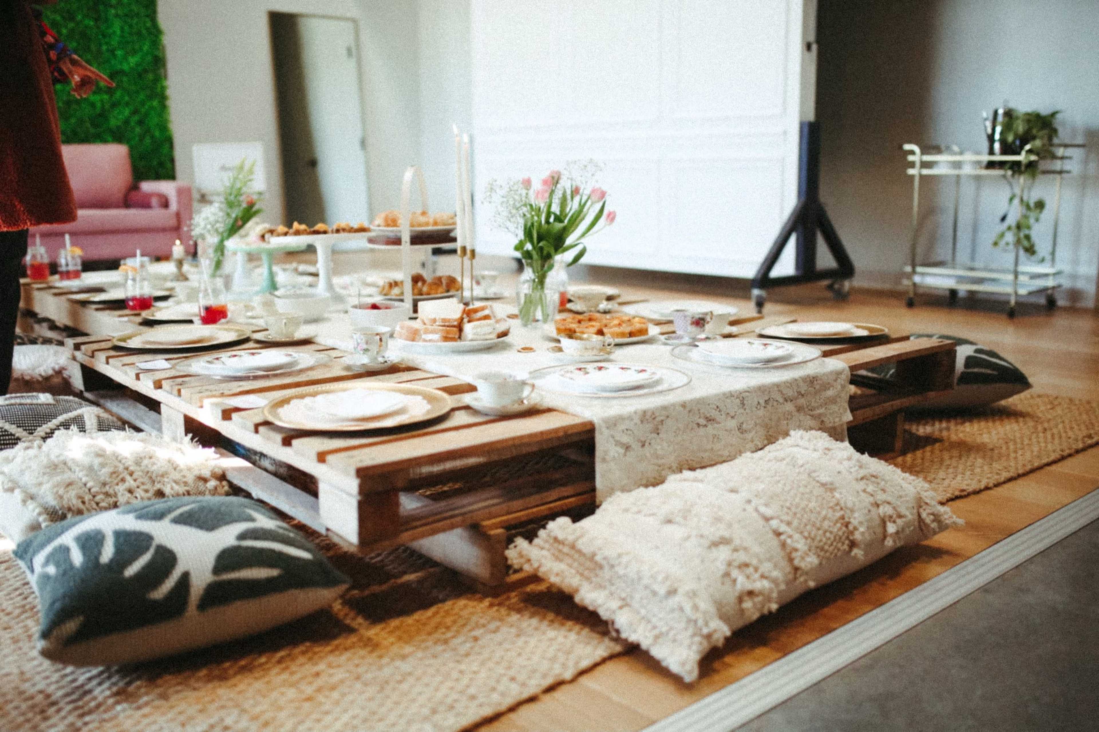 A low wooden table is set with tea cups and various snacks, surrounded by cushions on a woven rug in an indoor space.