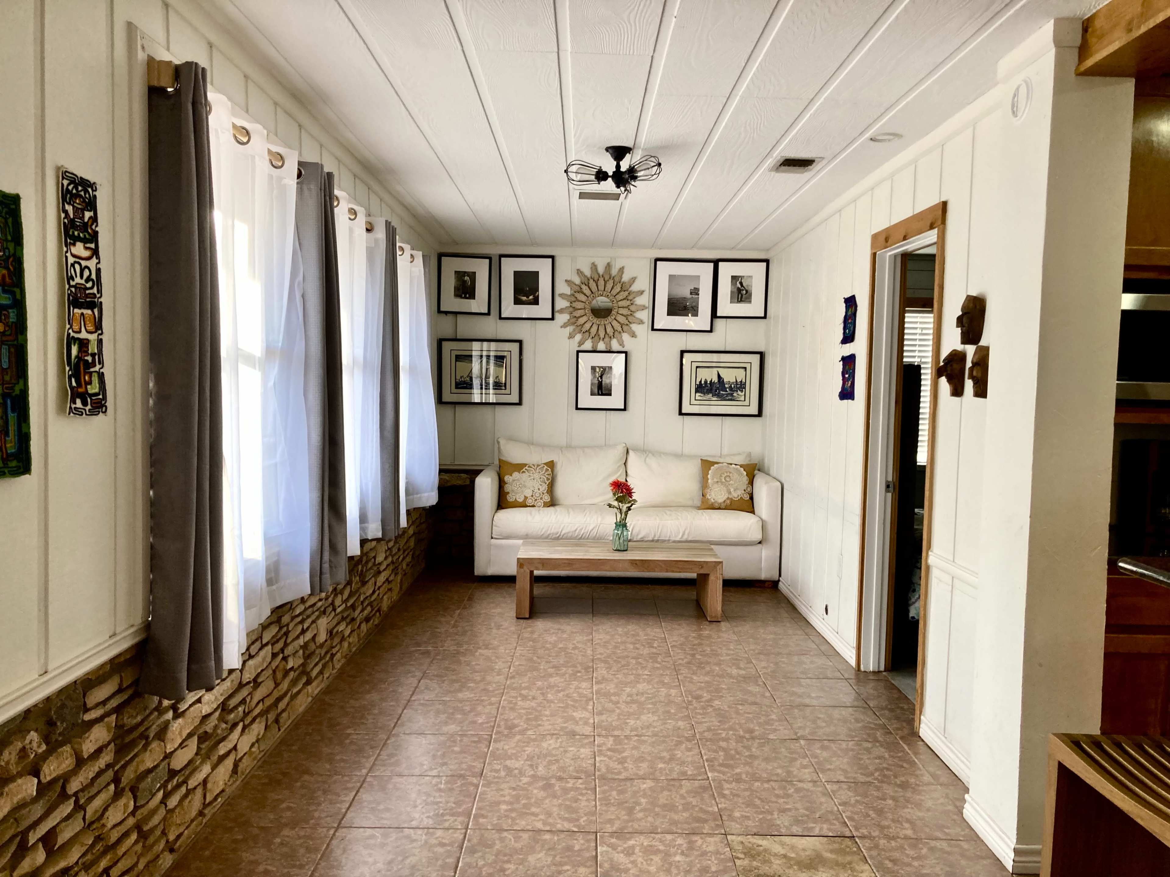 A living room featuring a white couch with decorative pillows, a wooden coffee table, and framed pictures on the wall, accompanied by large windows with curtains.