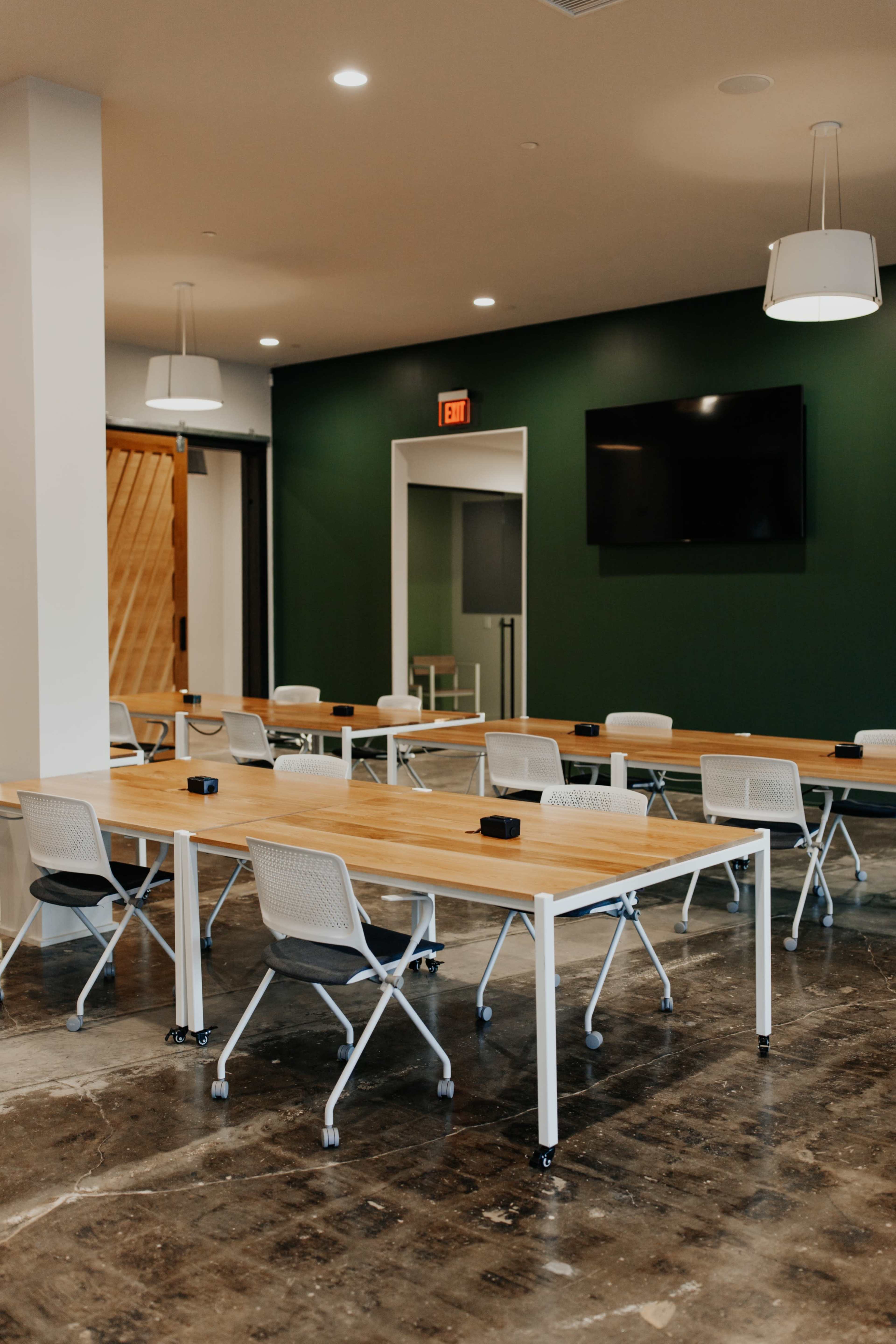A modern meeting room features a series of long tables with wheels and chairs arranged in an open layout, complemented by a dark green accent wall and a television screen.