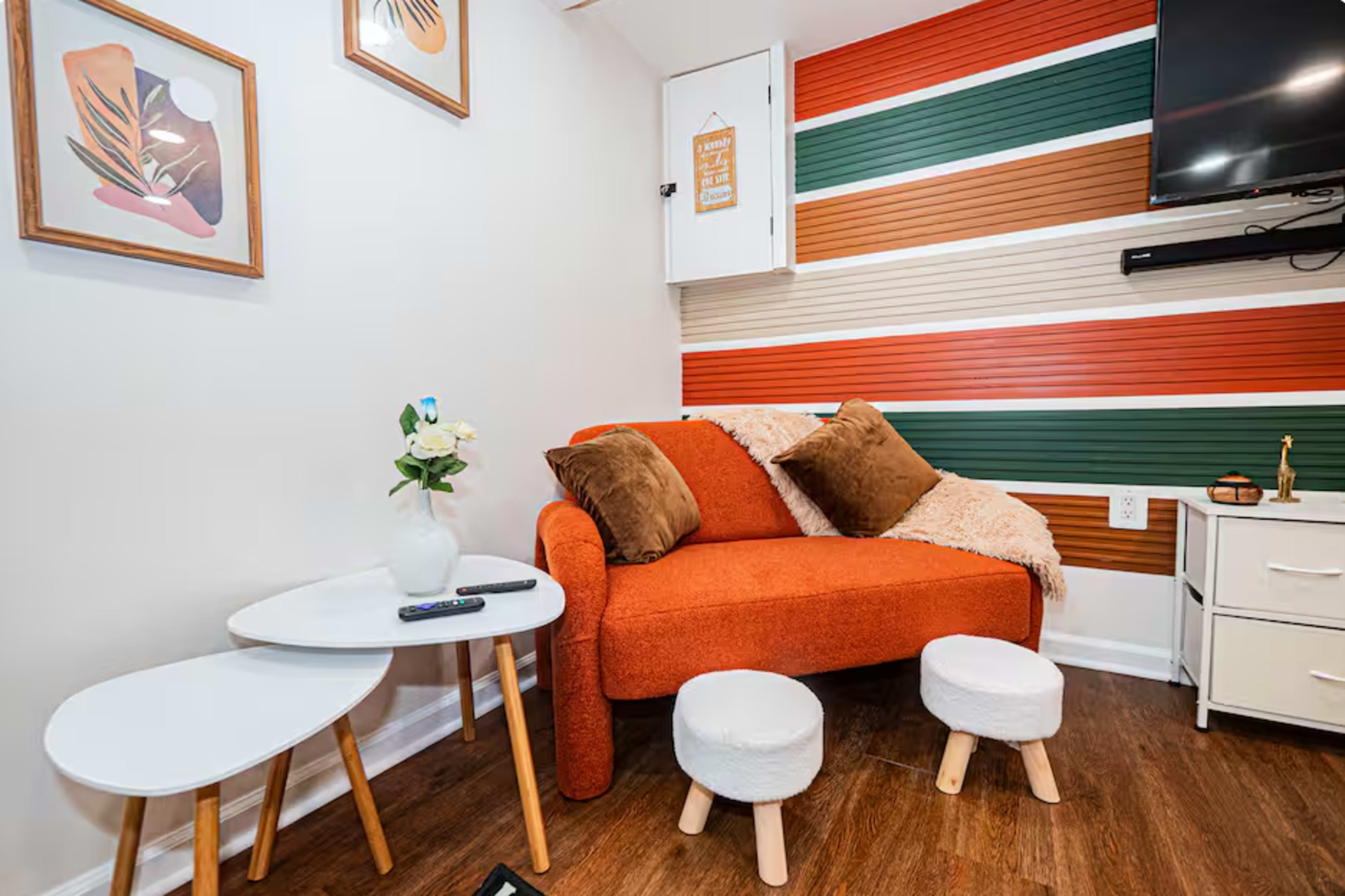 The image shows a cozy seating area with an orange couch, two small white stools, and a decorative wall featuring horizontal stripes in earthy tones.