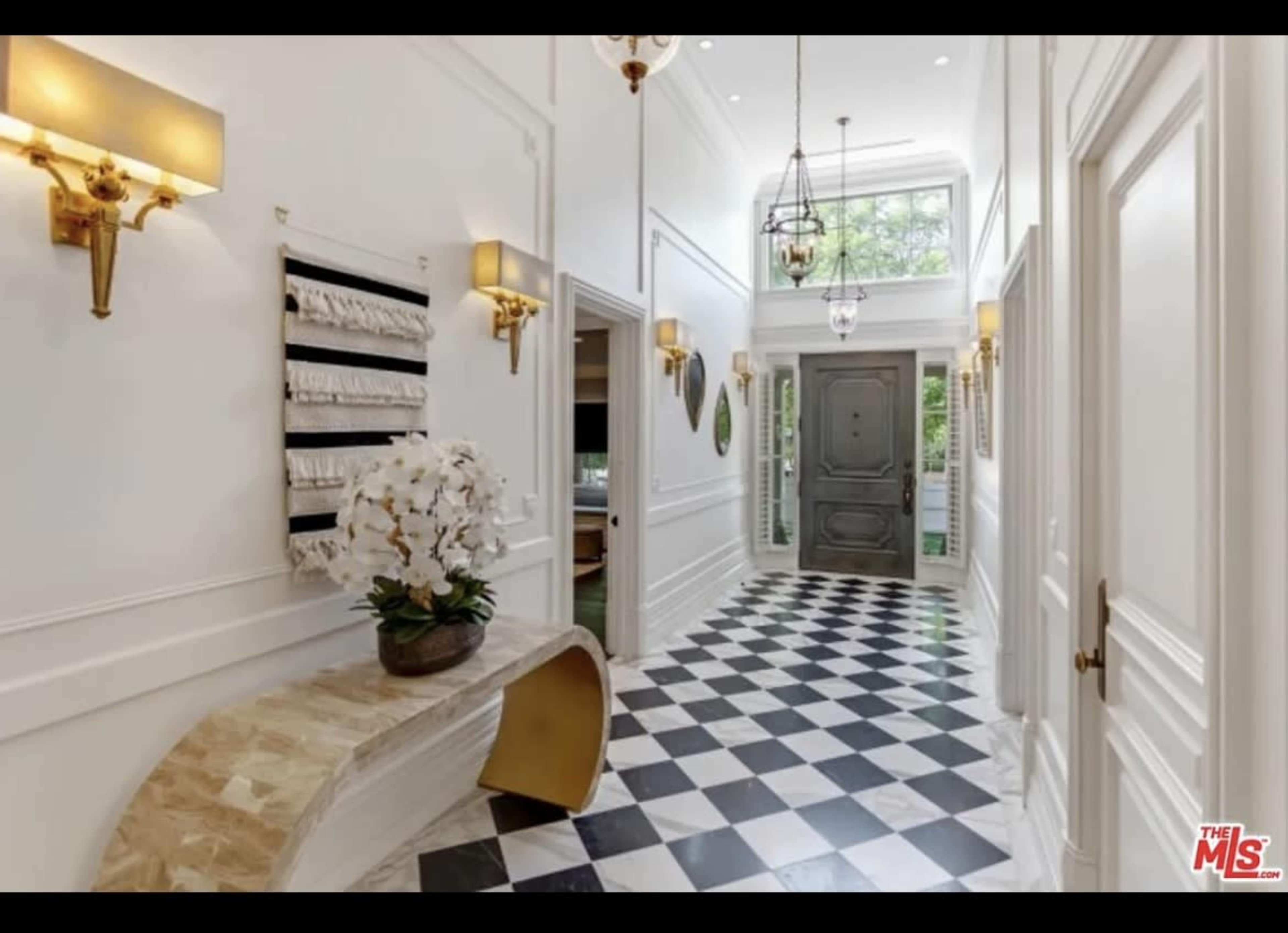 A well-lit hallway featuring a checkered black and white floor, ornate wall sconces, and a decorative console table with a floral arrangement.