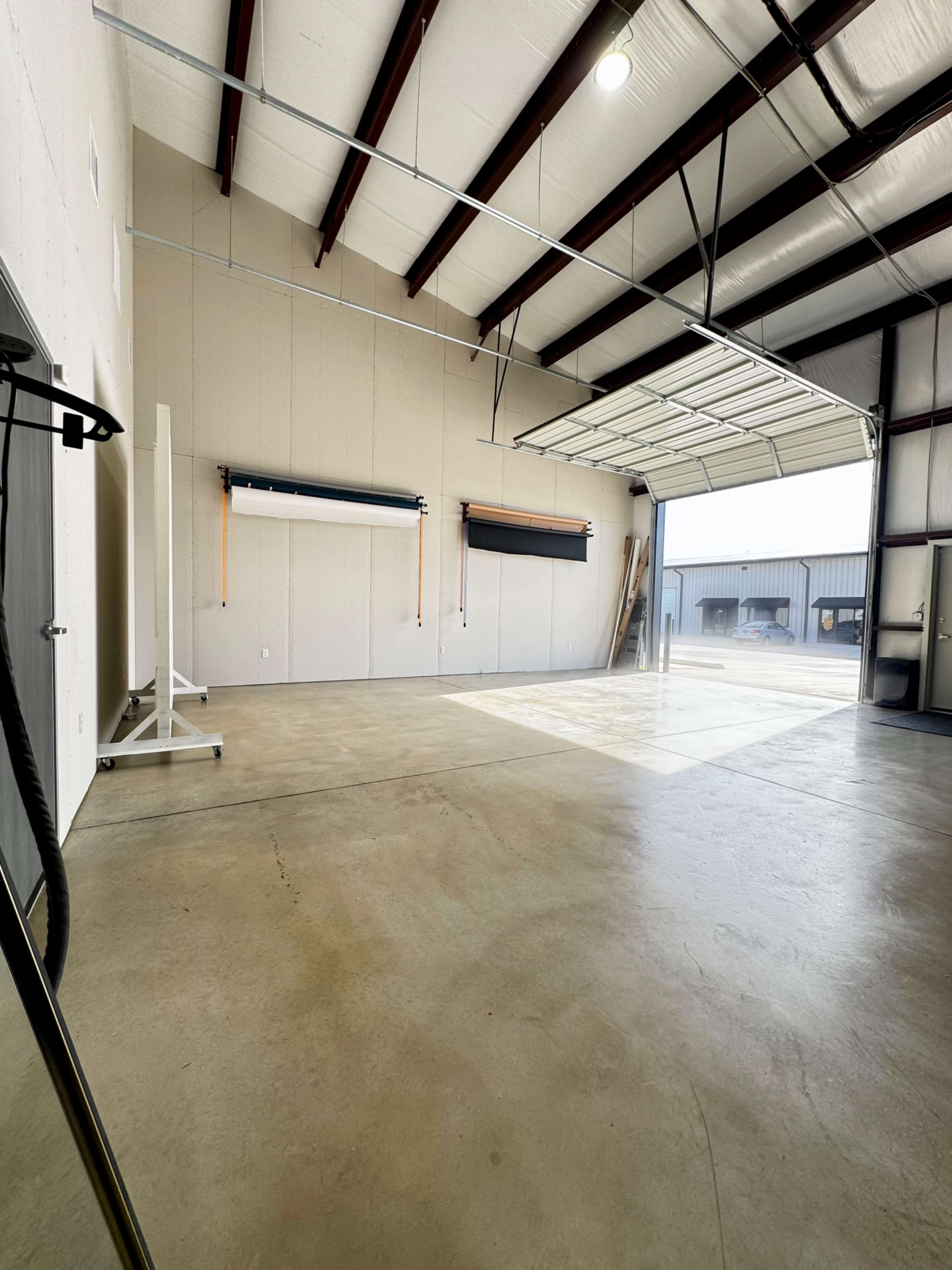 The image shows an empty industrial garage with a concrete floor, high metal beams, and a large overhead door partially open, leading to an exterior area.
