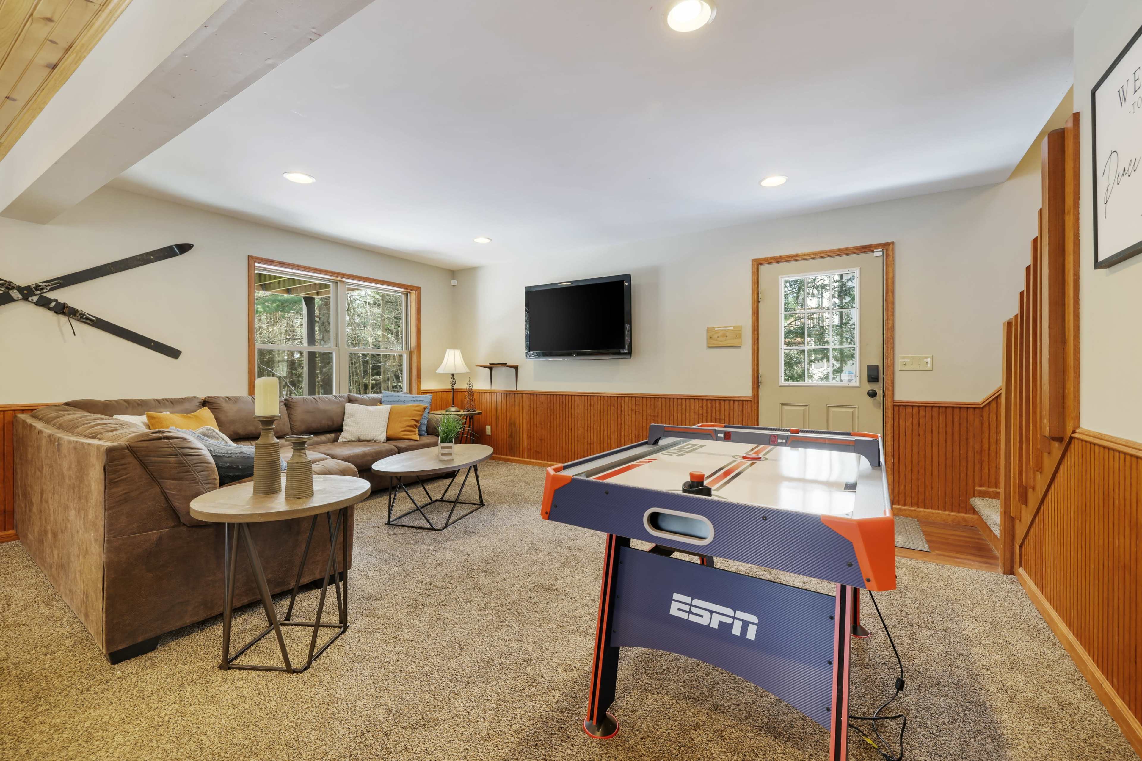 A cozy living room featuring a sectional sofa, a small table, and an air hockey table, with a television mounted on the wall and a door leading outside.