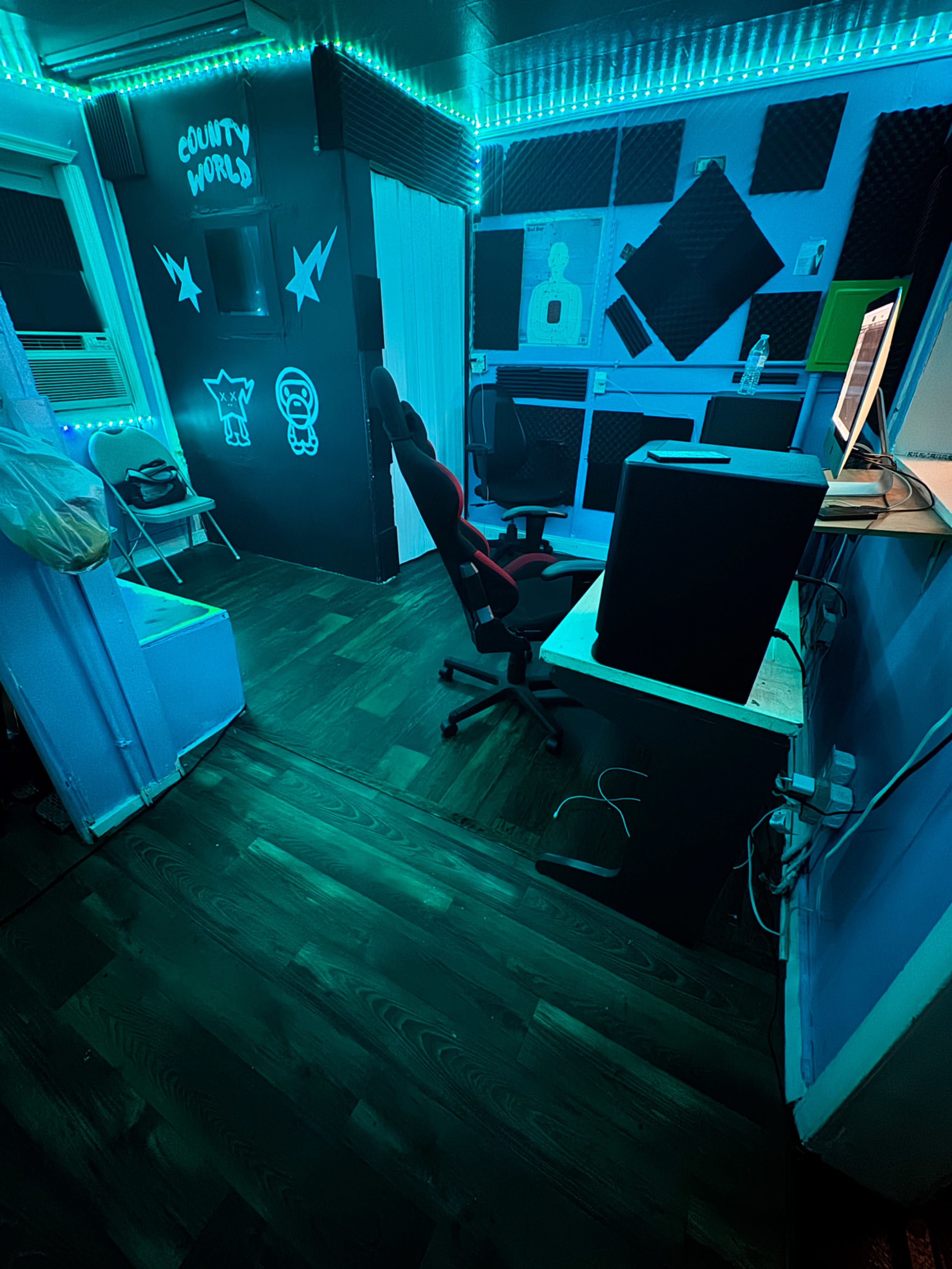 The image shows a compact recording studio with blue LED lighting, acoustic panels on the walls, and a desk with a computer and office chair.