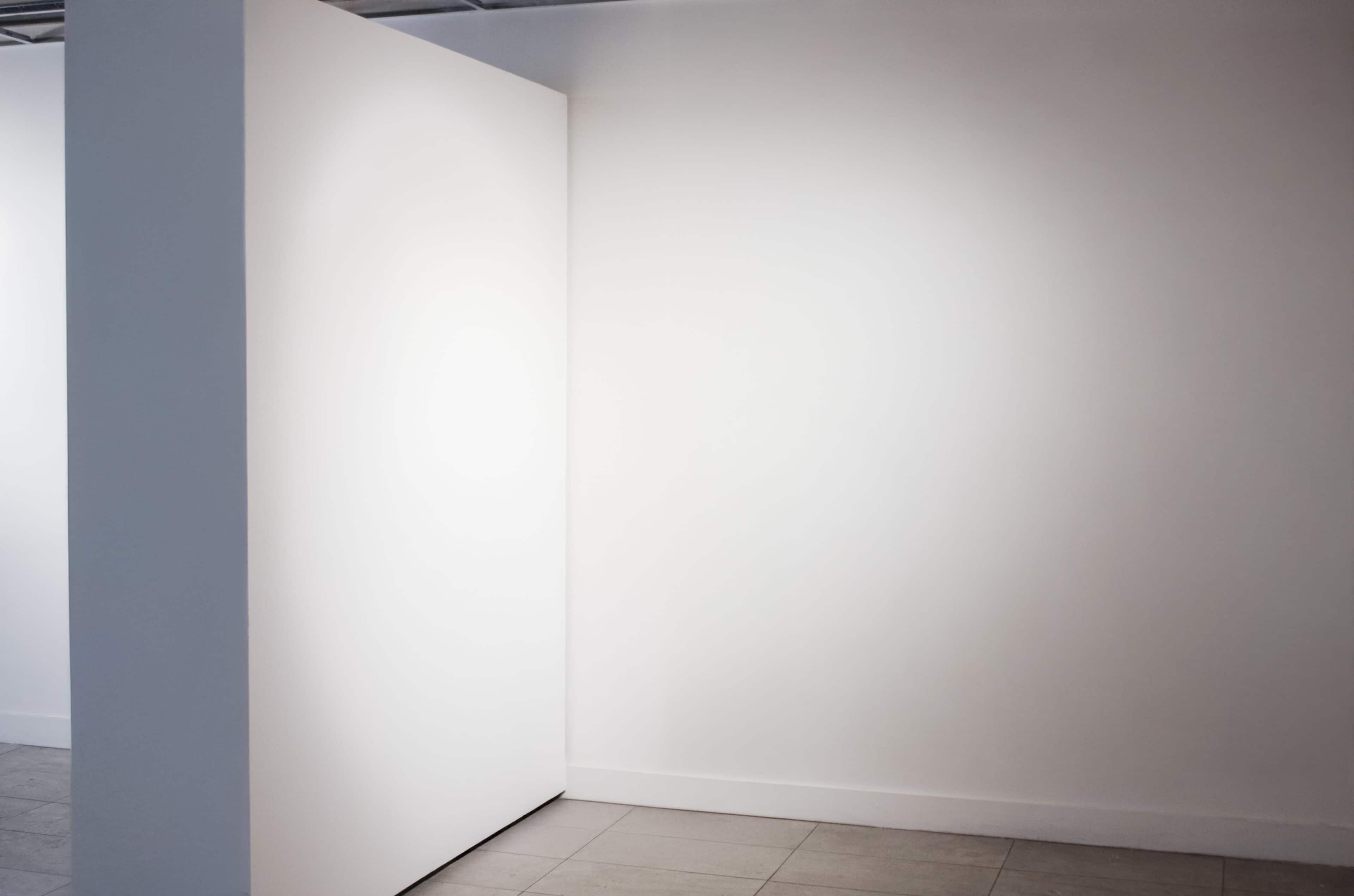 The image shows a corner of a minimalist gallery with two white walls and a tiled floor.