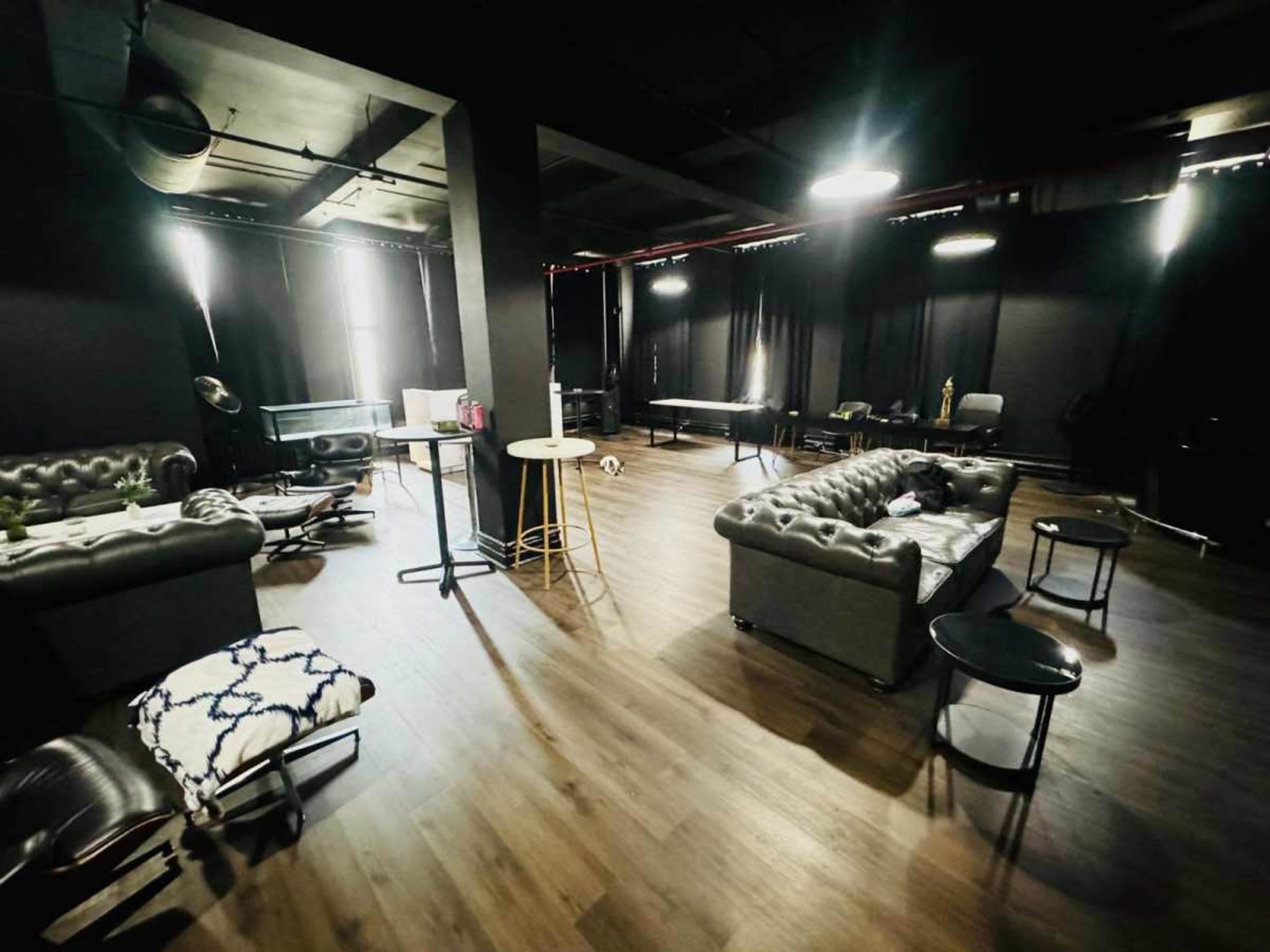 The image shows a spacious, dimly lit room with dark walls, featuring a mix of modern furniture, including couches, tables, and chairs.