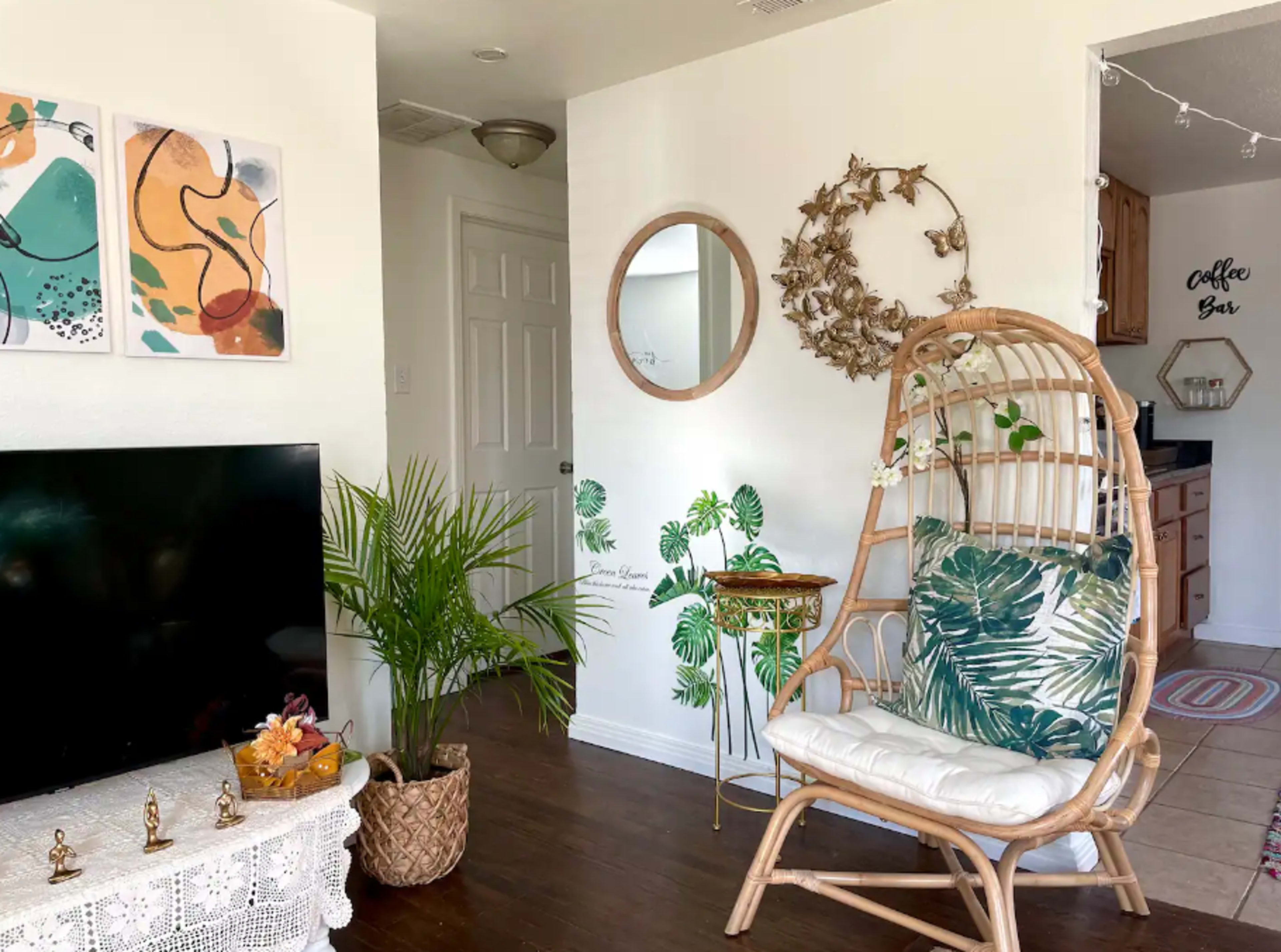 The scene depicts a cozy living space featuring a rattan chair with patterned cushions, a large TV, houseplants, wall art, and a circular mirror.