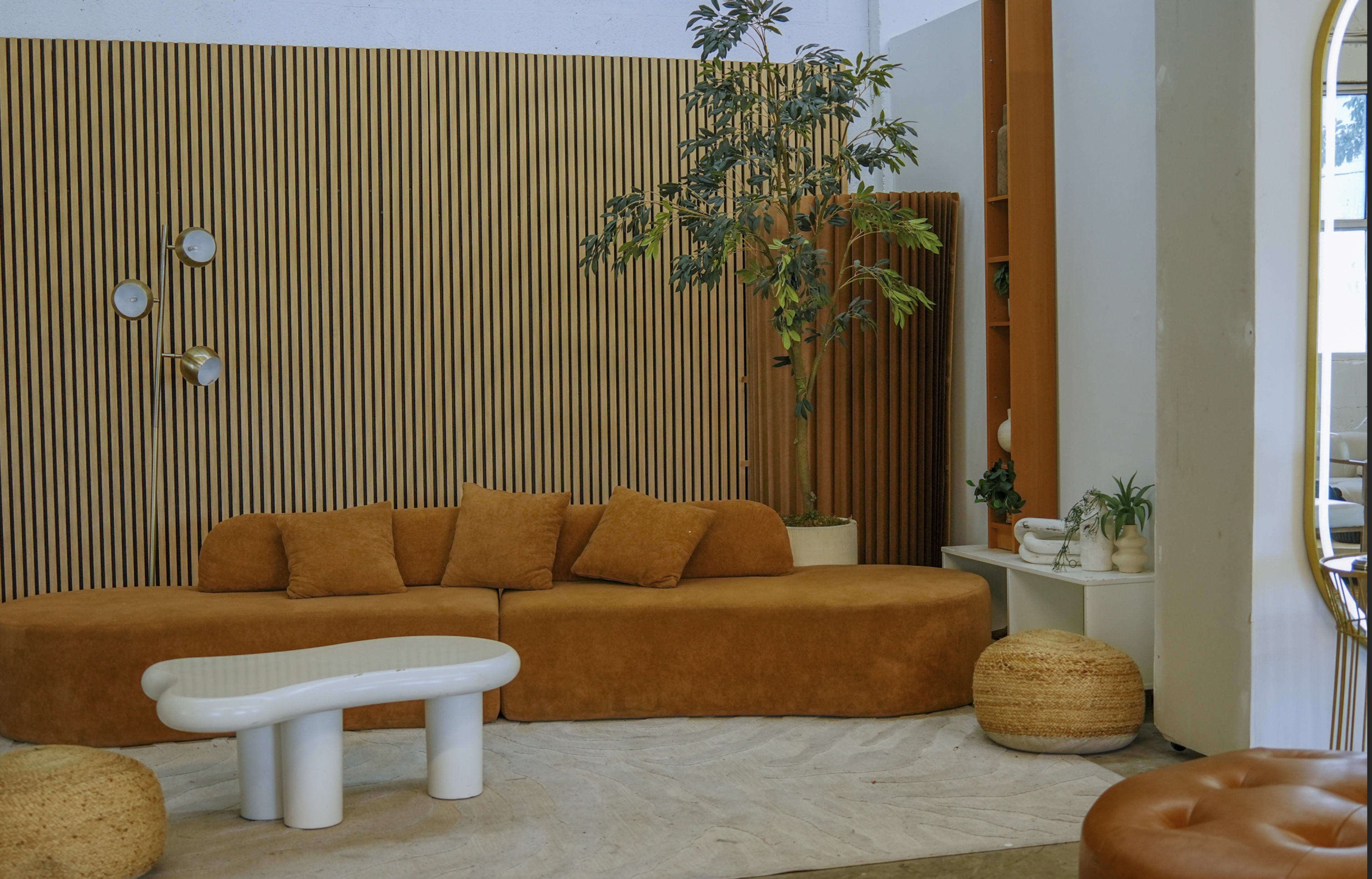 A curved brown sofa is positioned against a wooden slat wall, accompanied by a white table and potted plants in a modern living space.