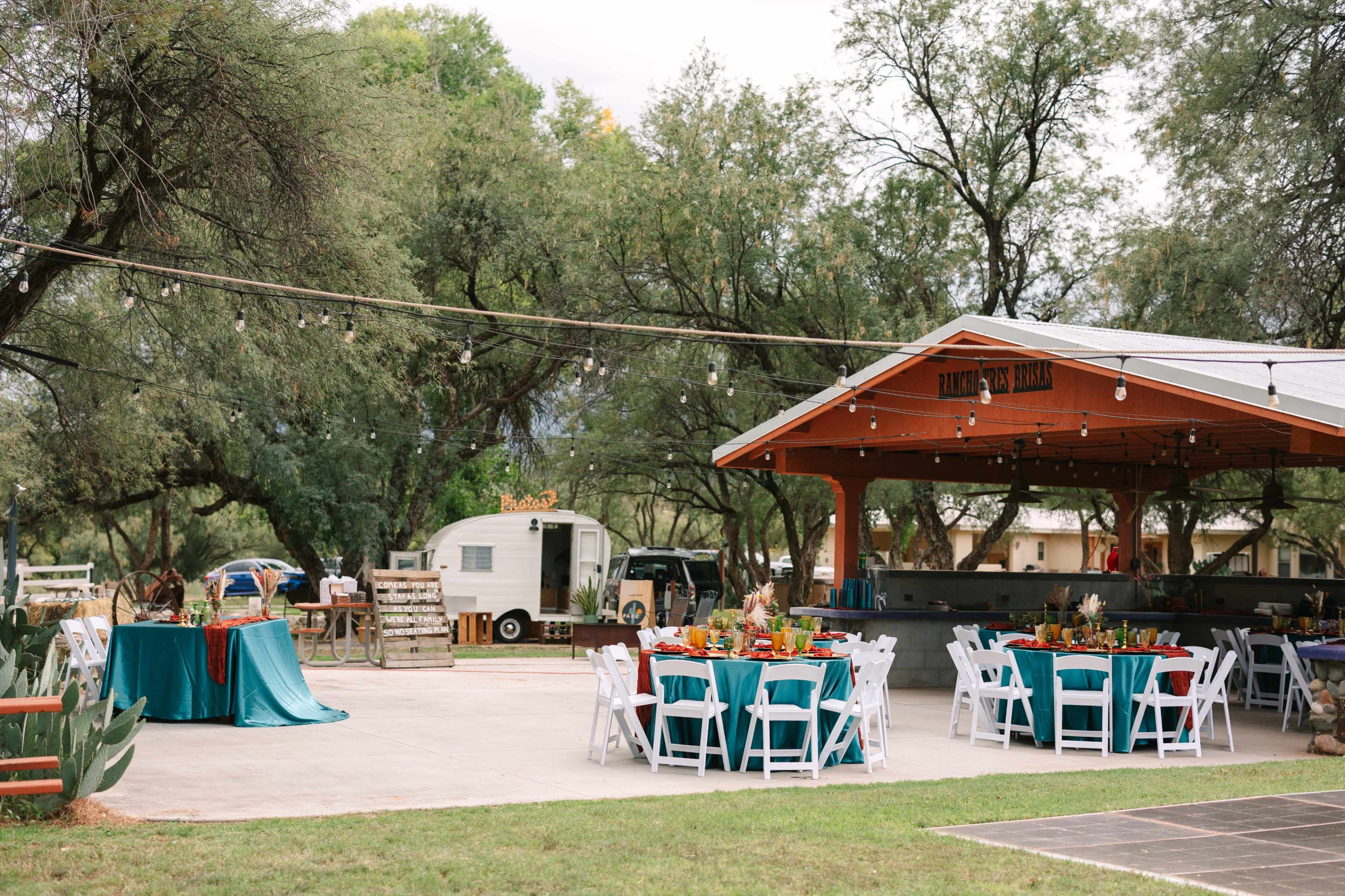 The outdoor venue features round tables with teal tablecloths set up under a covered area, surrounded by greenery and string lights, with a vintage trailer visible in the background.