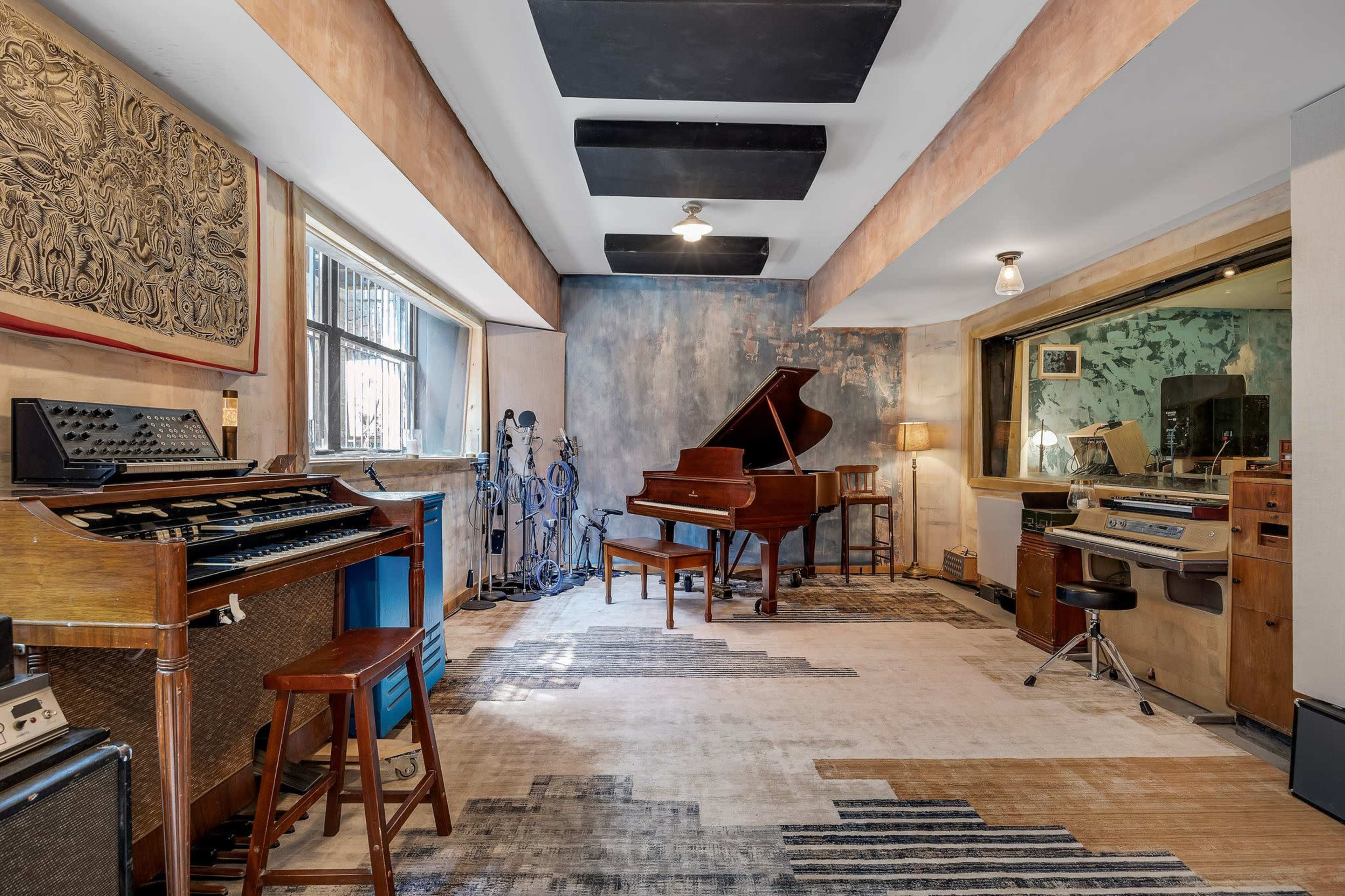 A spacious music studio featuring a grand piano, vintage electronic instruments, microphone stands, and large windows.