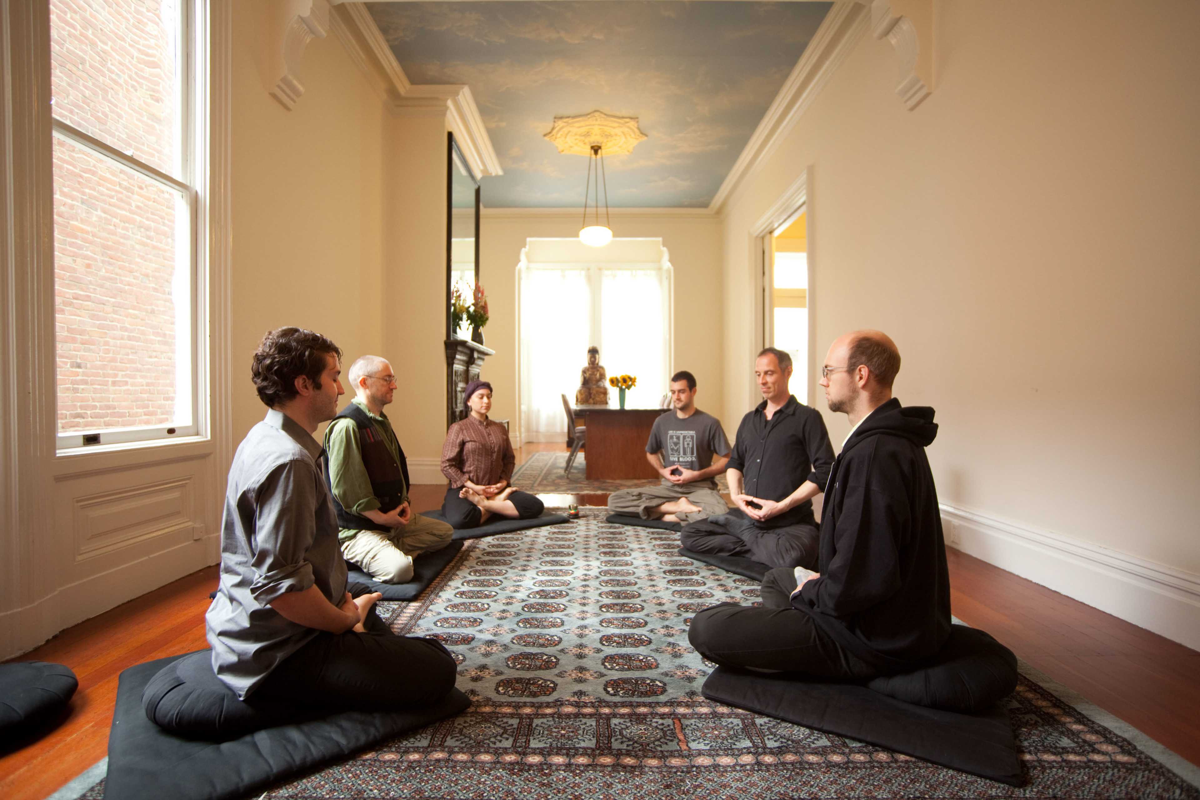 A group of six individuals sits cross-legged on floor cushions in a spacious room with a decorative rug, while a statue of a seated figure and a woman are observed in the background.