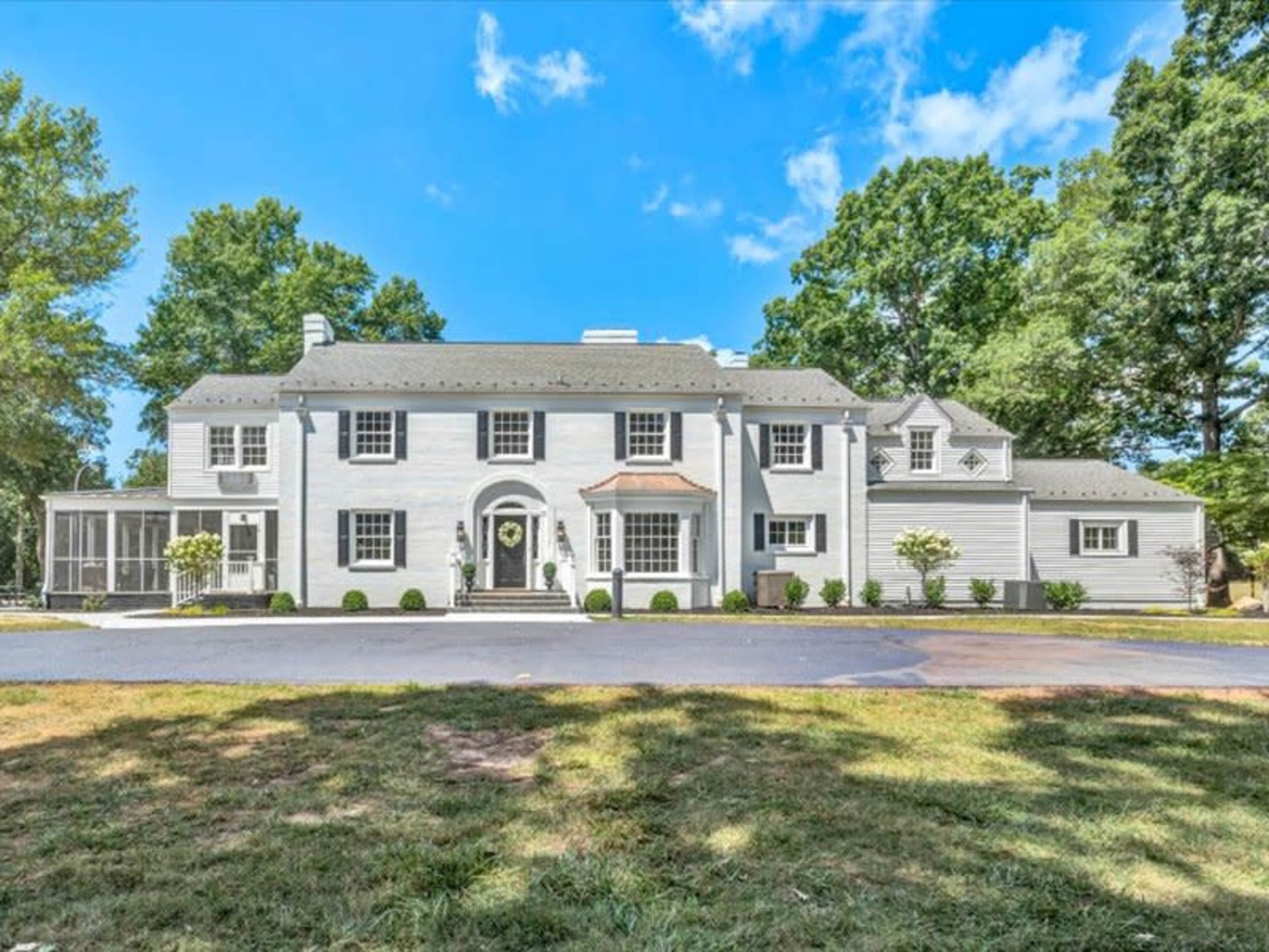 The image shows a large white two-story house with a manicured lawn and several trees surrounding it.