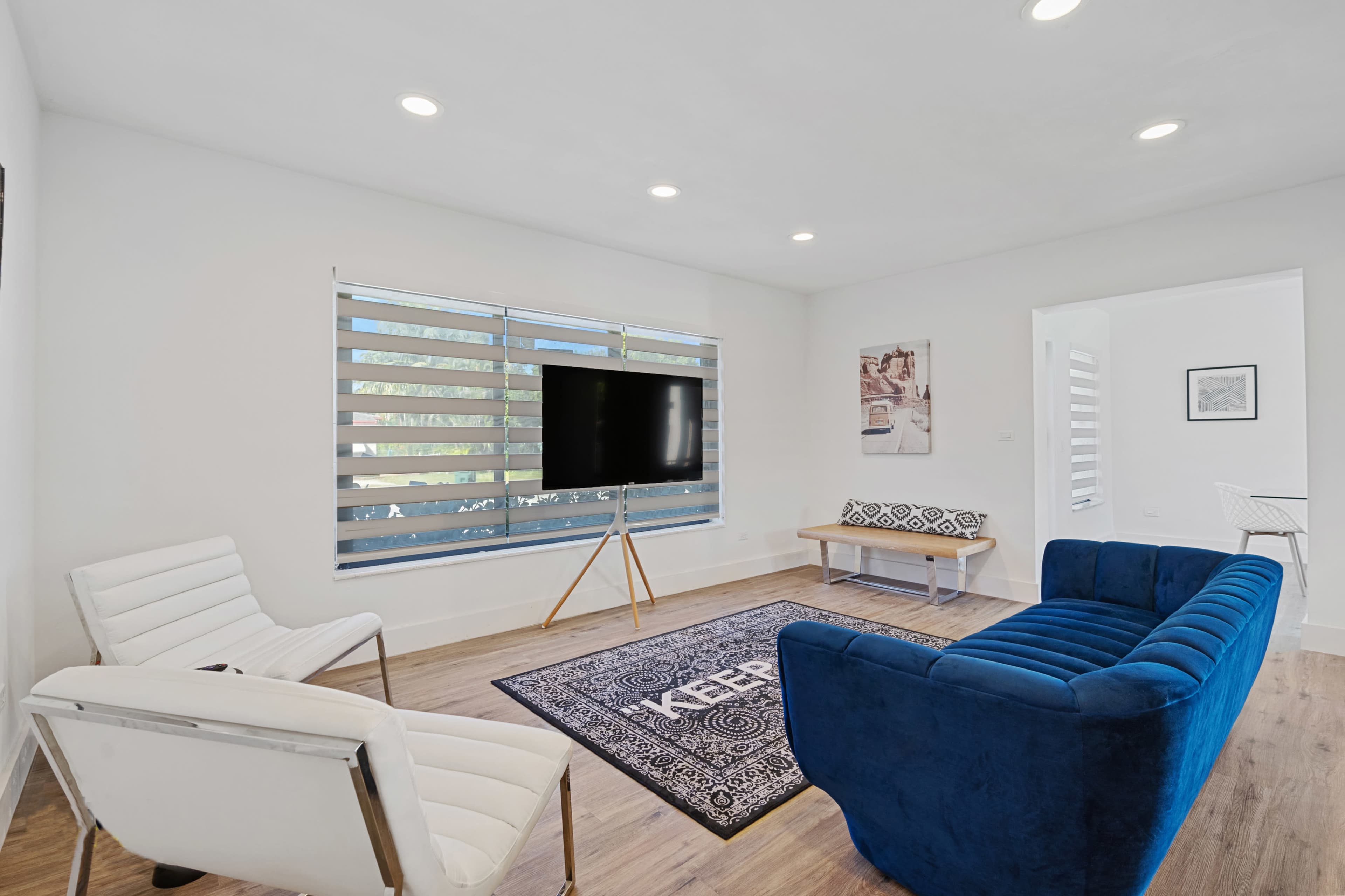 The image shows a modern living room with a blue couch, white chairs, a flat-screen TV on a stand, and a patterned rug on the wooden floor.