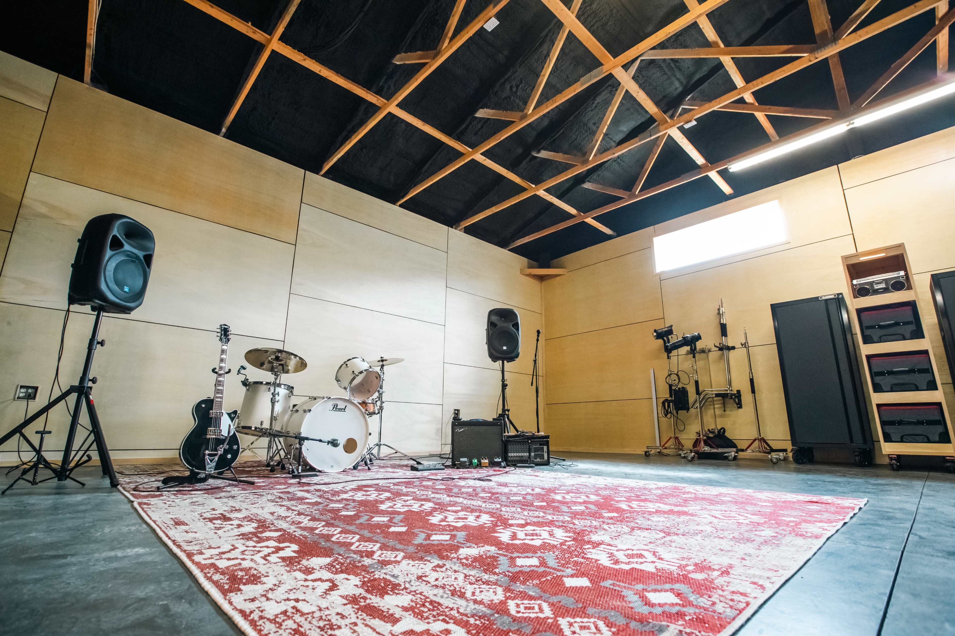 The image shows a music rehearsal space featuring a drum set, amplifiers, a guitar, and a patterned rug on a concrete floor.