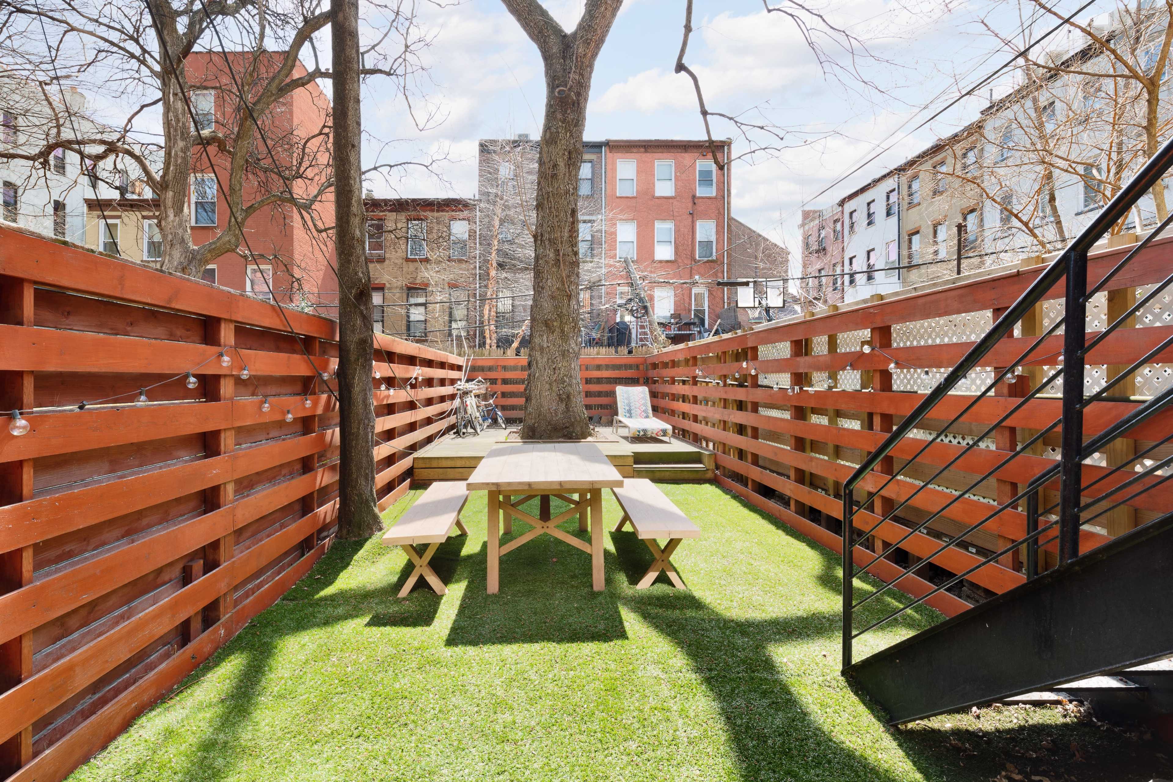 A fenced backyard with a wooden picnic table on grass, surrounded by buildings and trees.
