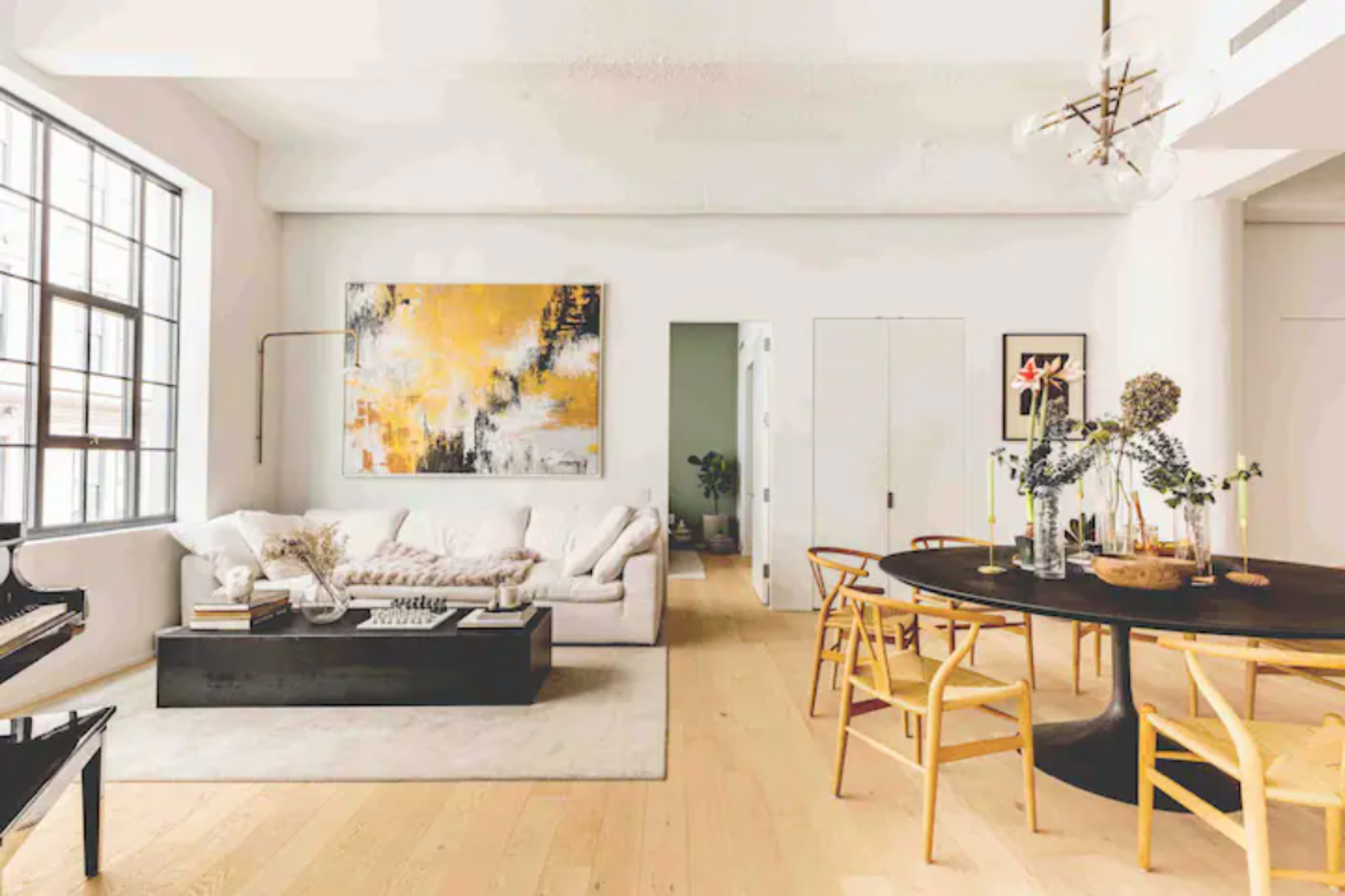 A modern living room features a light-colored sofa, a black coffee table, and a dining area with a round table surrounded by chairs, illuminated by large windows.