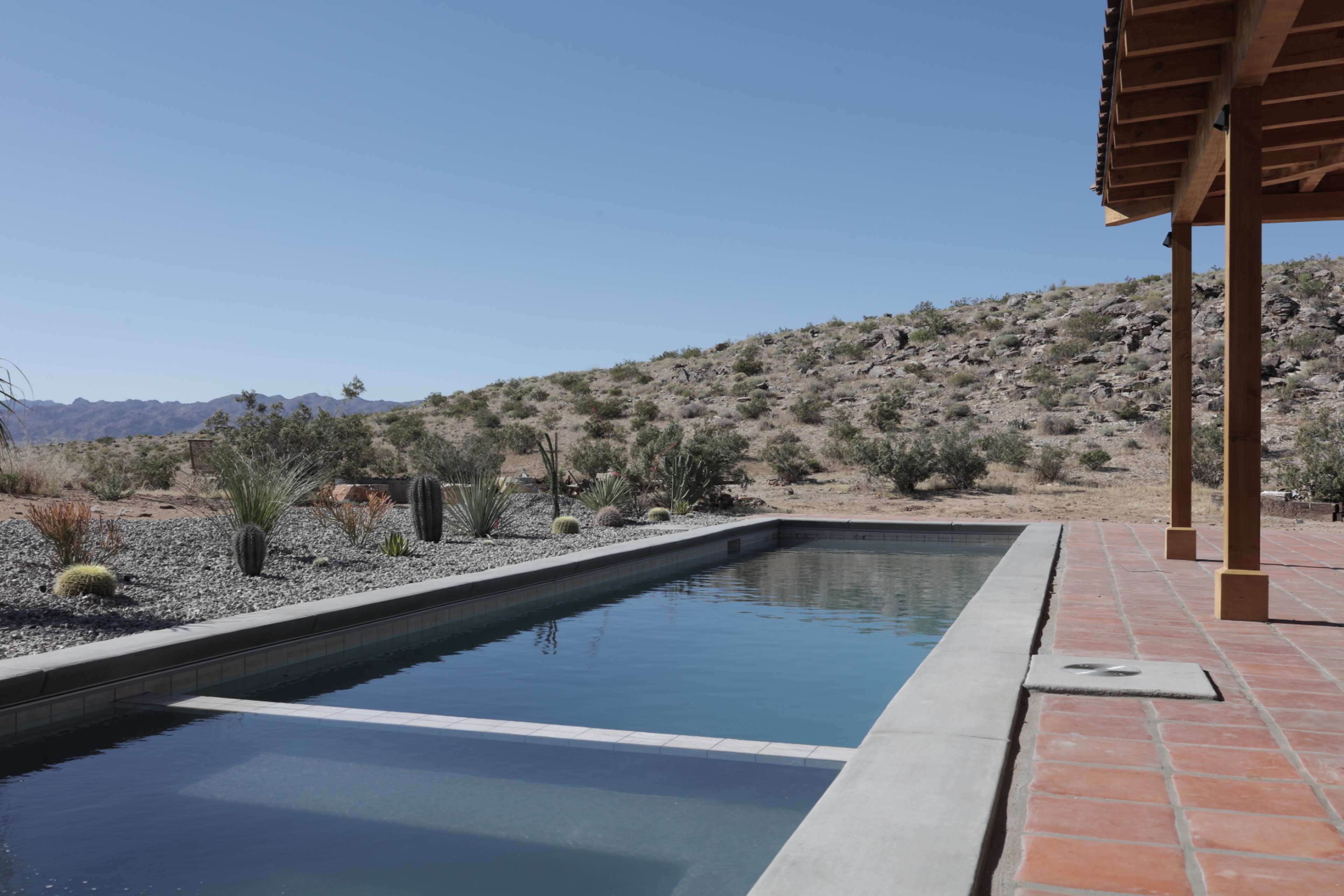 A rectangular swimming pool is situated next to a desert landscape featuring sparse vegetation and rocky hills under a clear blue sky.