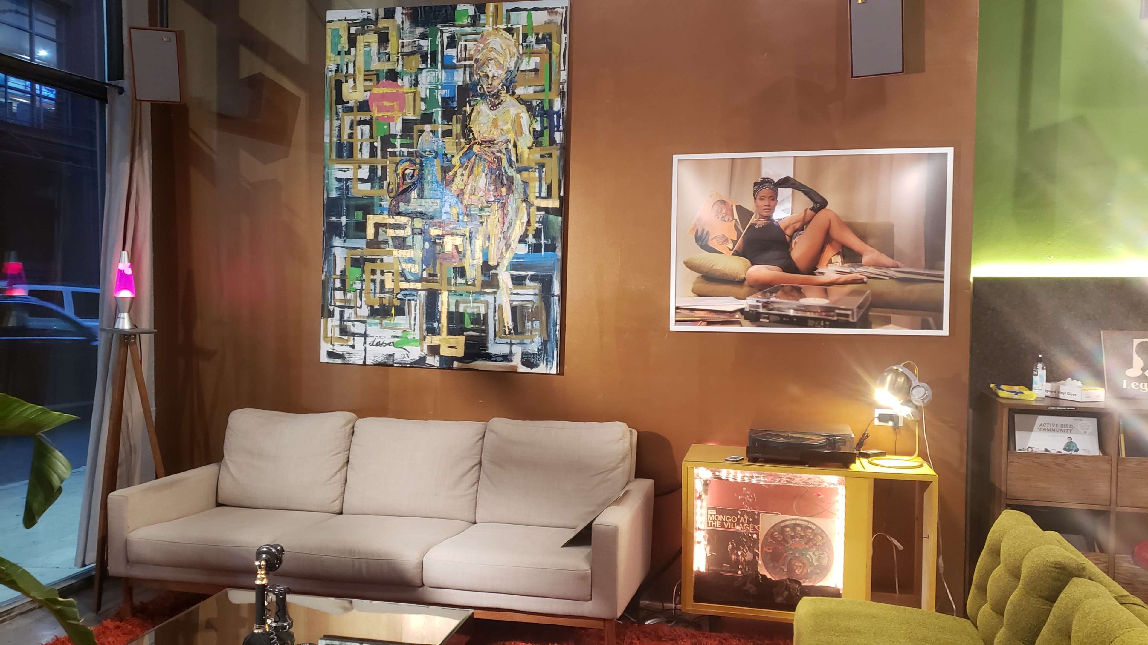 A modern interior featuring a beige couch, two framed artworks, a green chair, and a record player set against a brown wall.