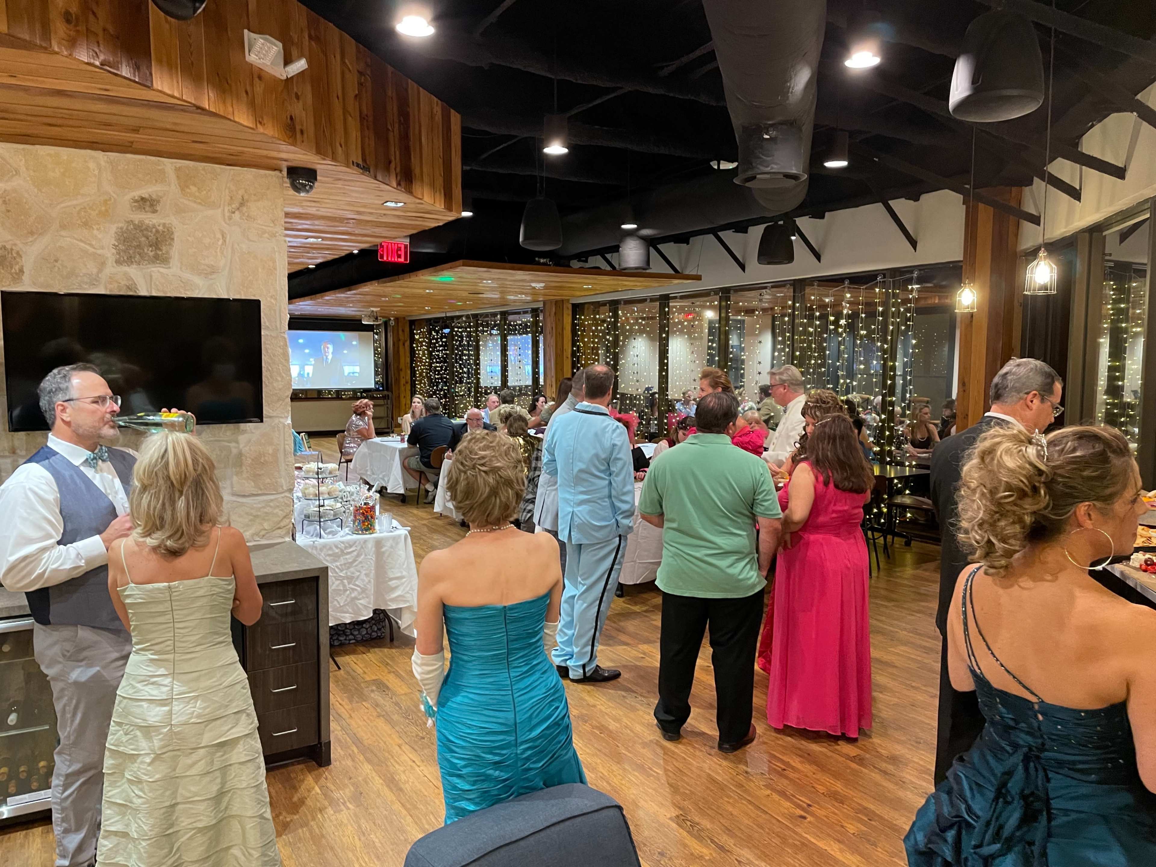 A group of attendees in formal attire socialize at a well-lit indoor event with a stone fireplace and string lights in the background.