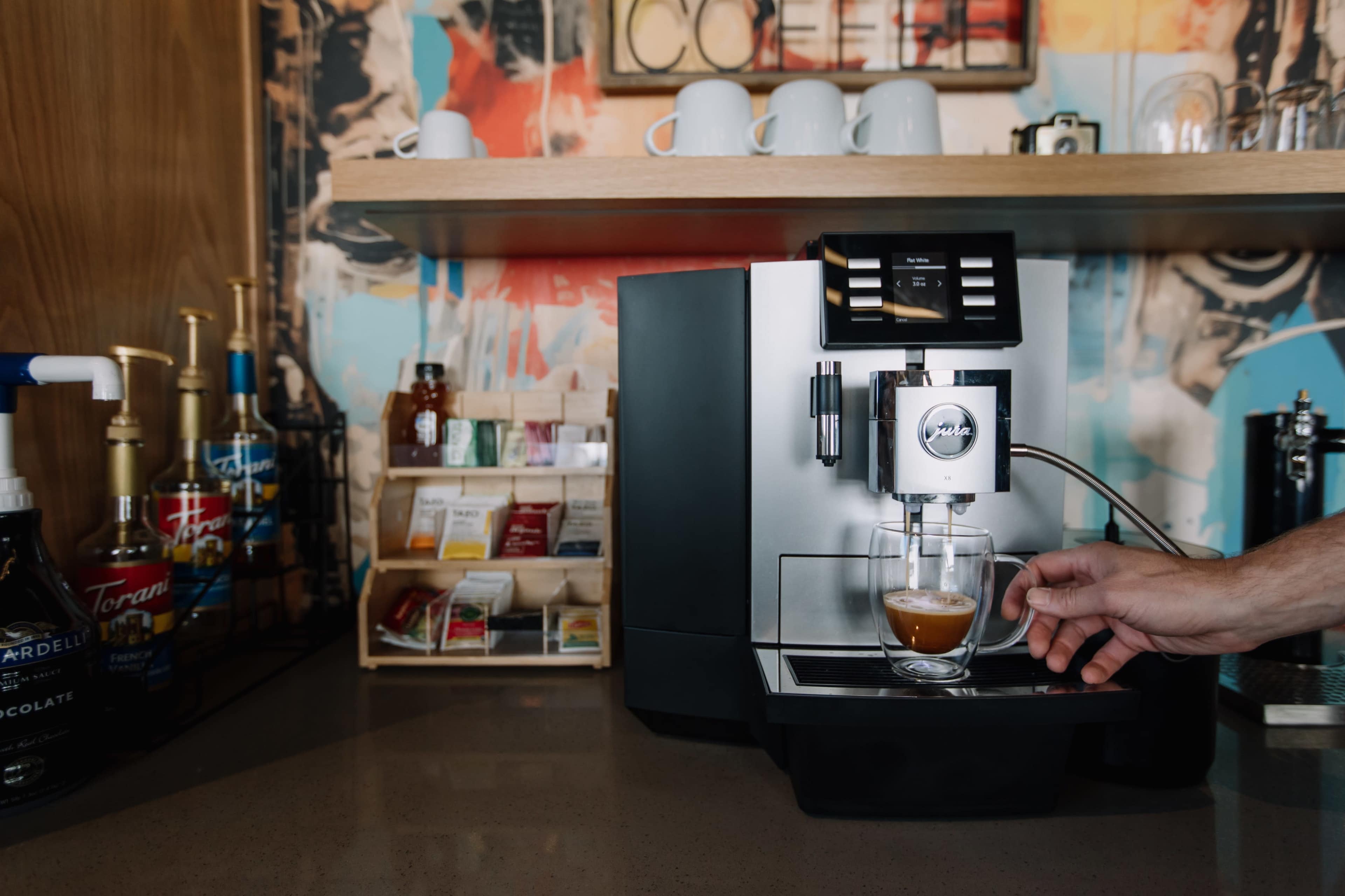 A hand is placing a glass under the spout of an espresso machine on a countertop, with shelves in the background displaying cups and various beverages.