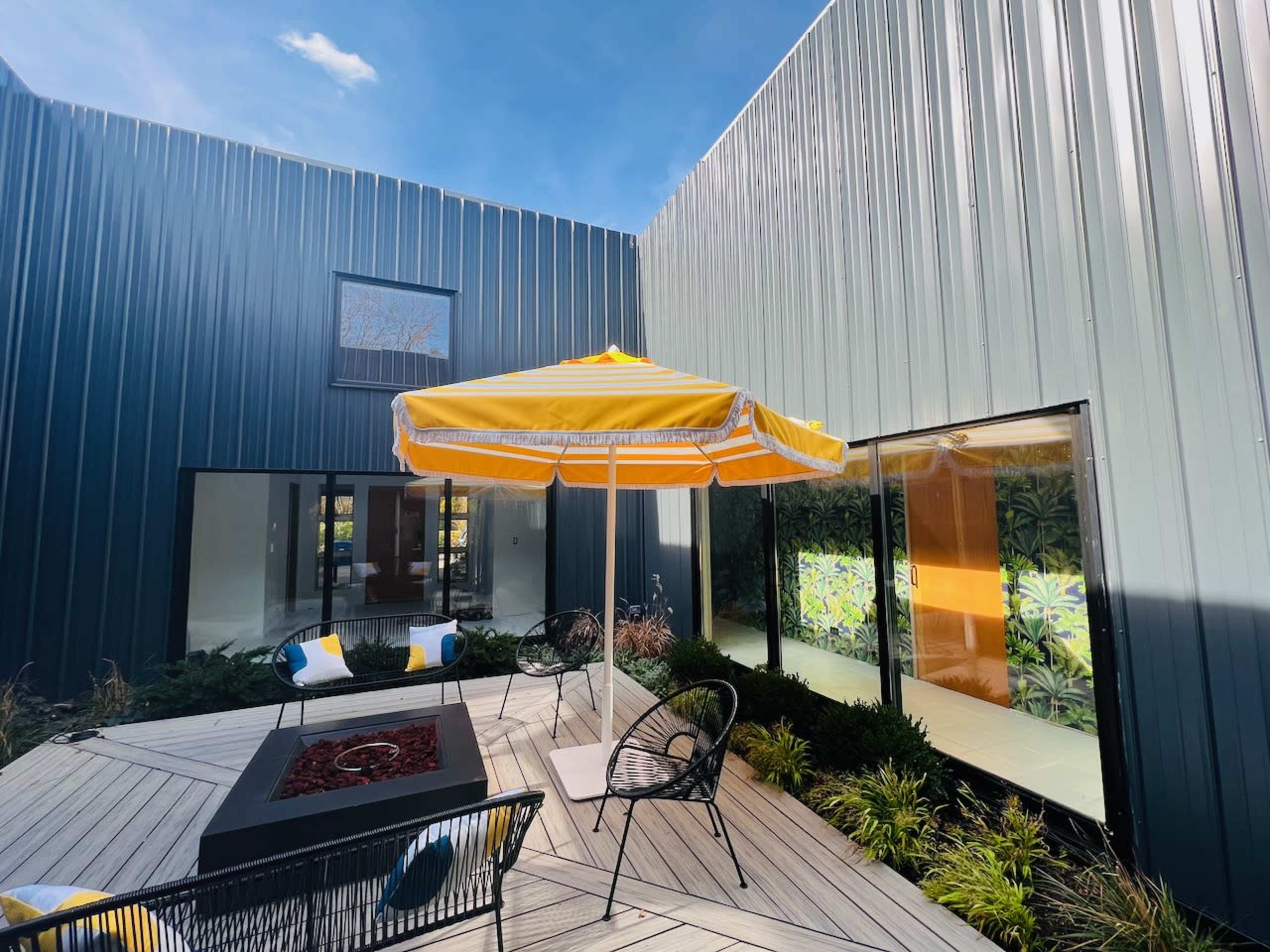 A modern courtyard with a yellow-striped umbrella, black chairs, and a fire pit, framed by two metal-clad buildings.