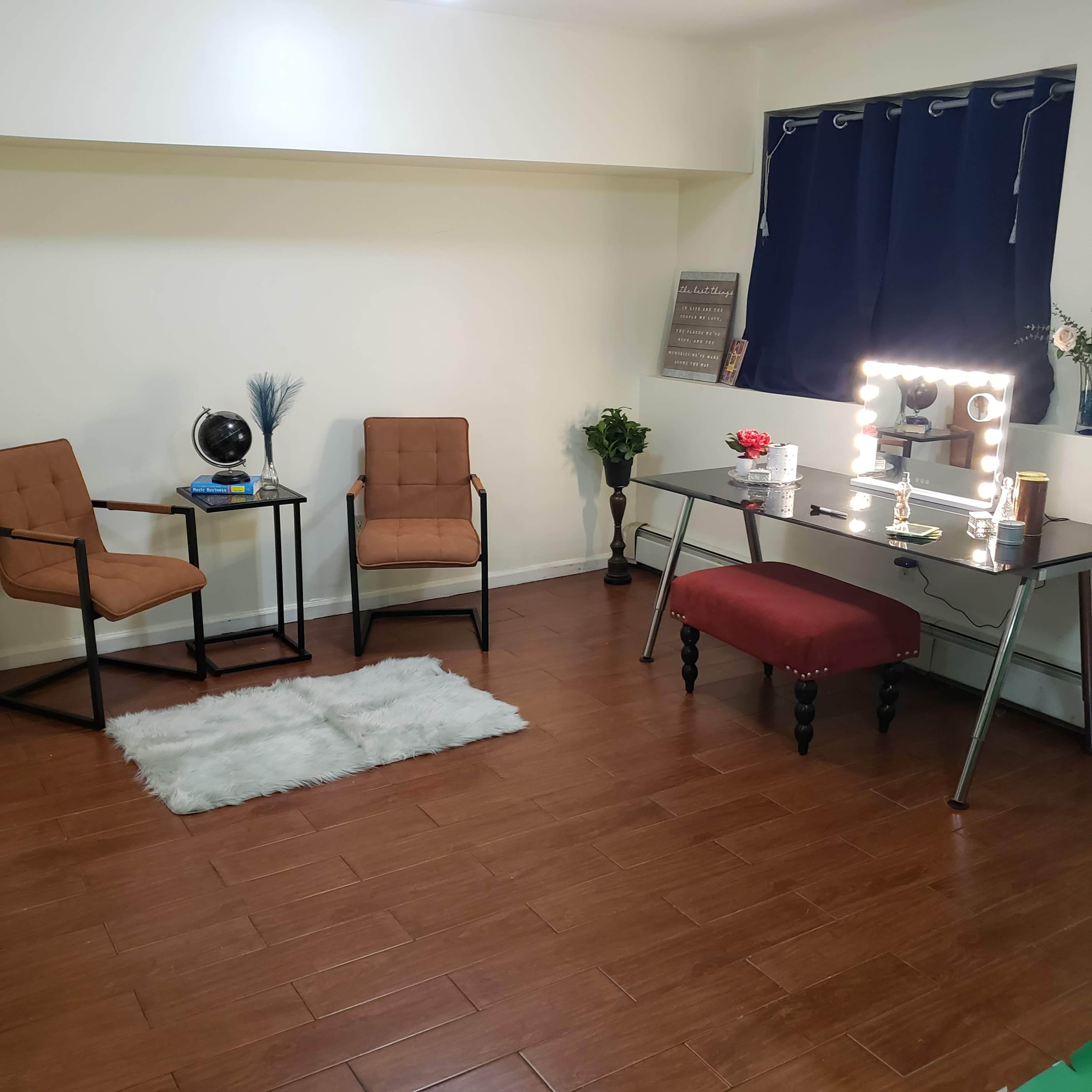 A minimalist room with two brown chairs, a vanity table featuring a large mirror and light bulbs, and a decorative accent rug on a wooden floor.