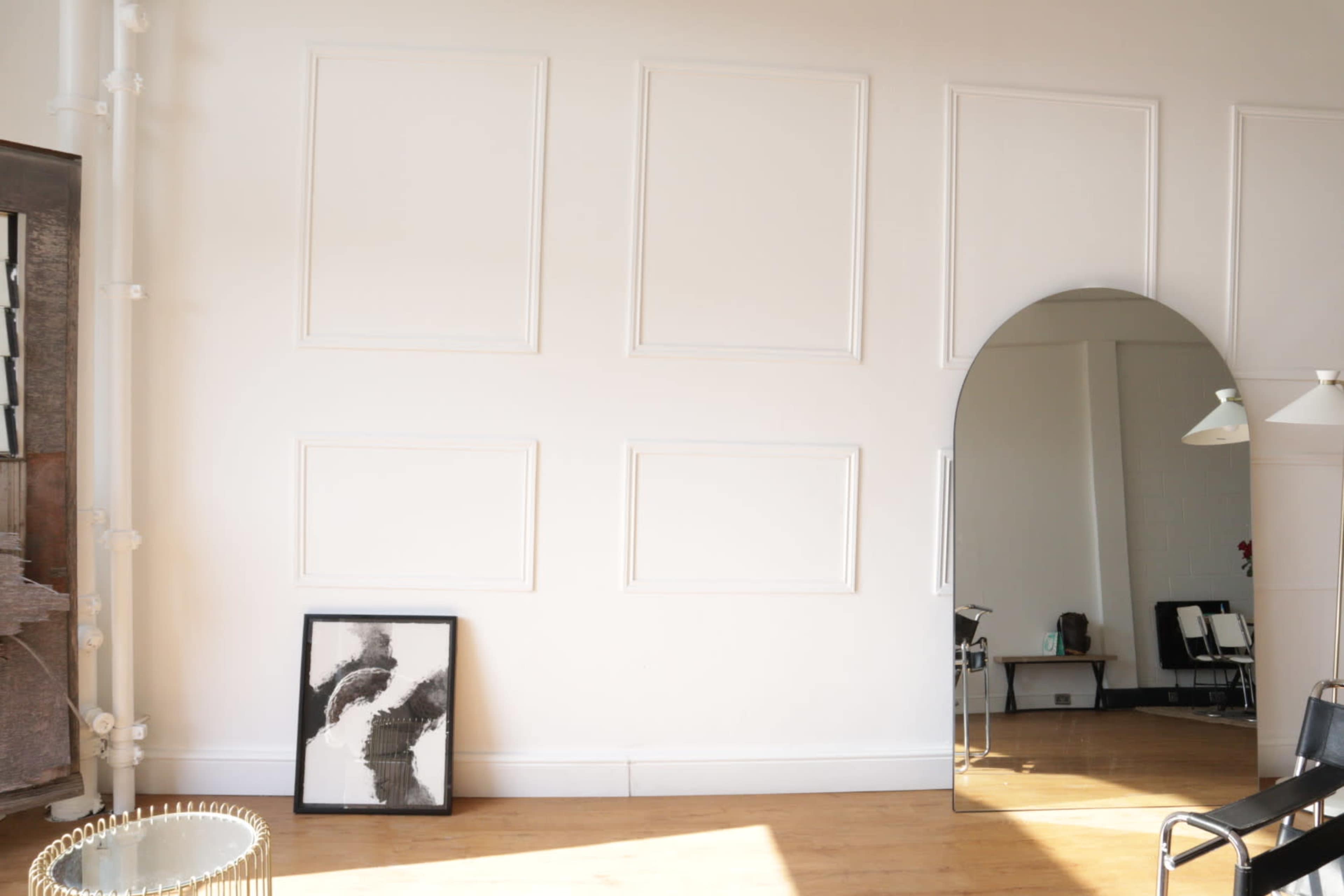 A bright, minimalist interior with white walls, wooden flooring, an arched mirror, and a framed artwork leaning against the wall.