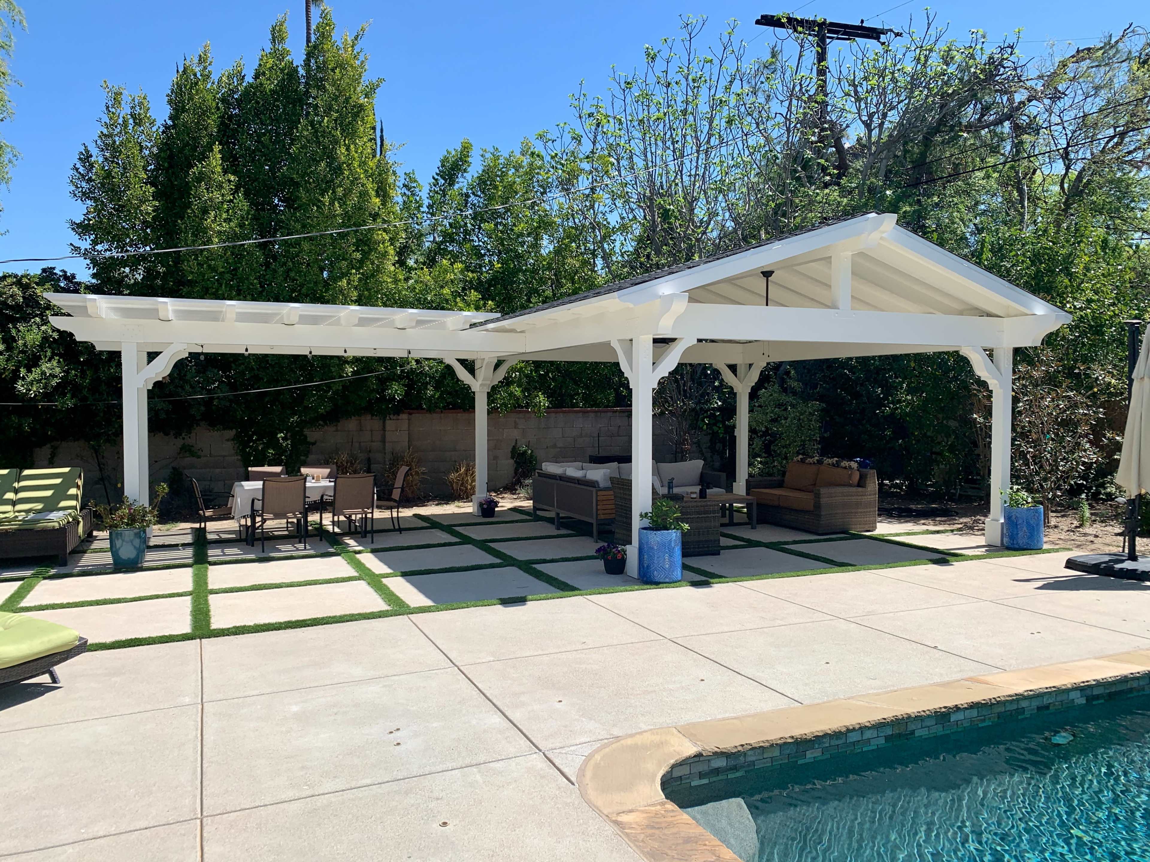 A shaded outdoor living space beside a swimming pool, featuring a white pergola, patio furniture, and lush greenery.
