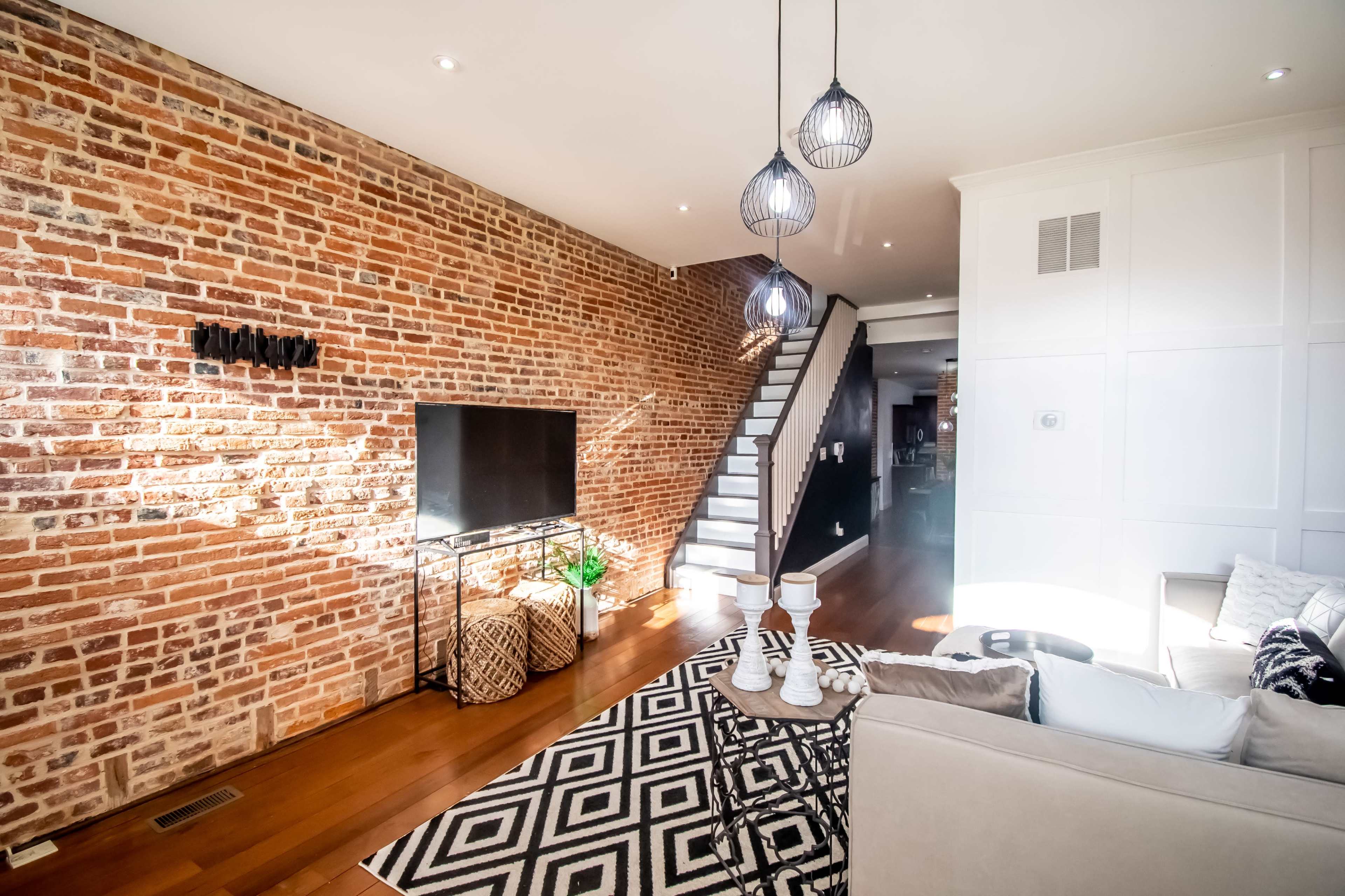 A living room with a brick accent wall, a staircase in the background, and modern lighting fixtures hanging above a stylish seating area.