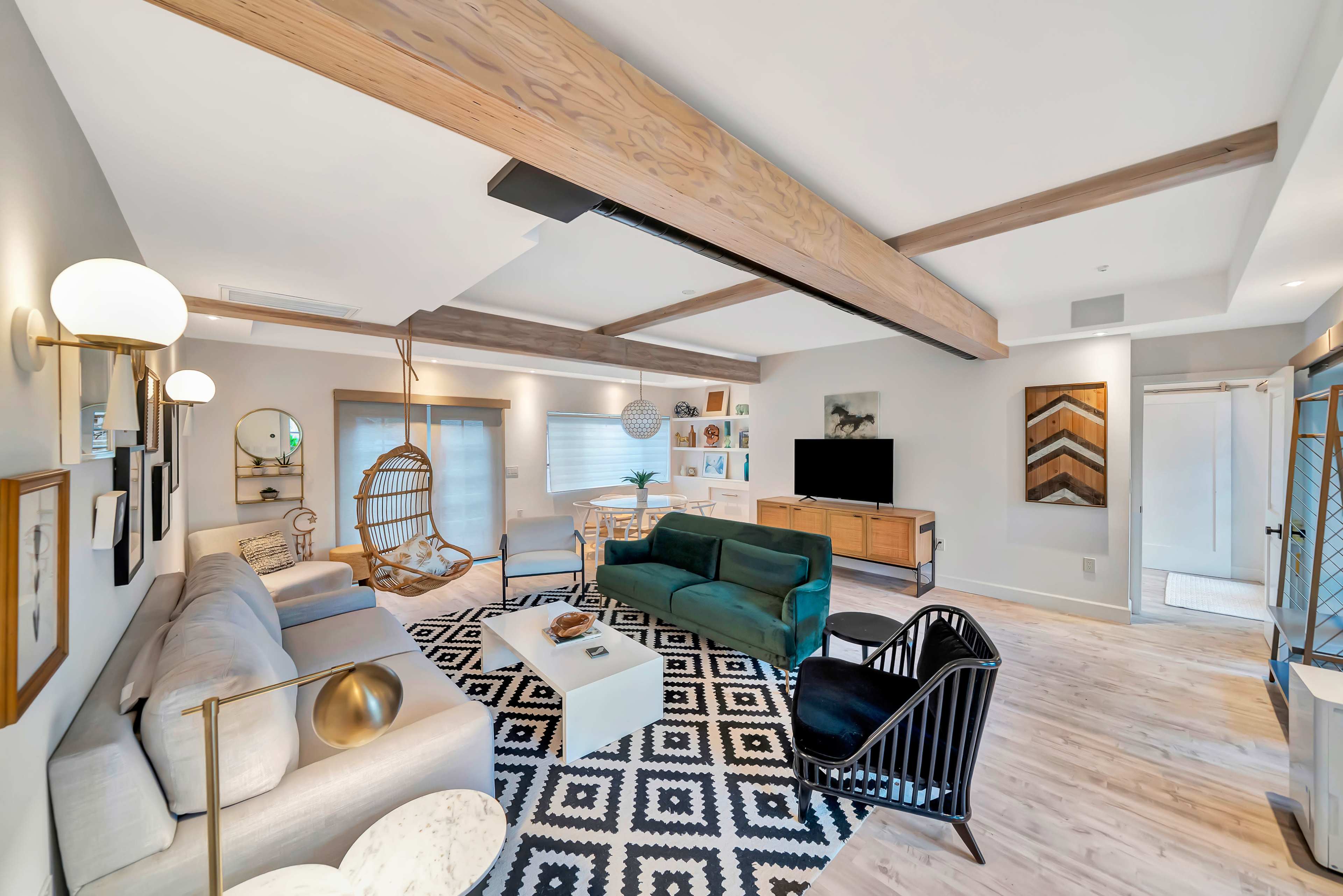 The image shows a modern living room with a combination of light-colored furniture, wooden beams on the ceiling, and a mix of decorative wall art and plants.