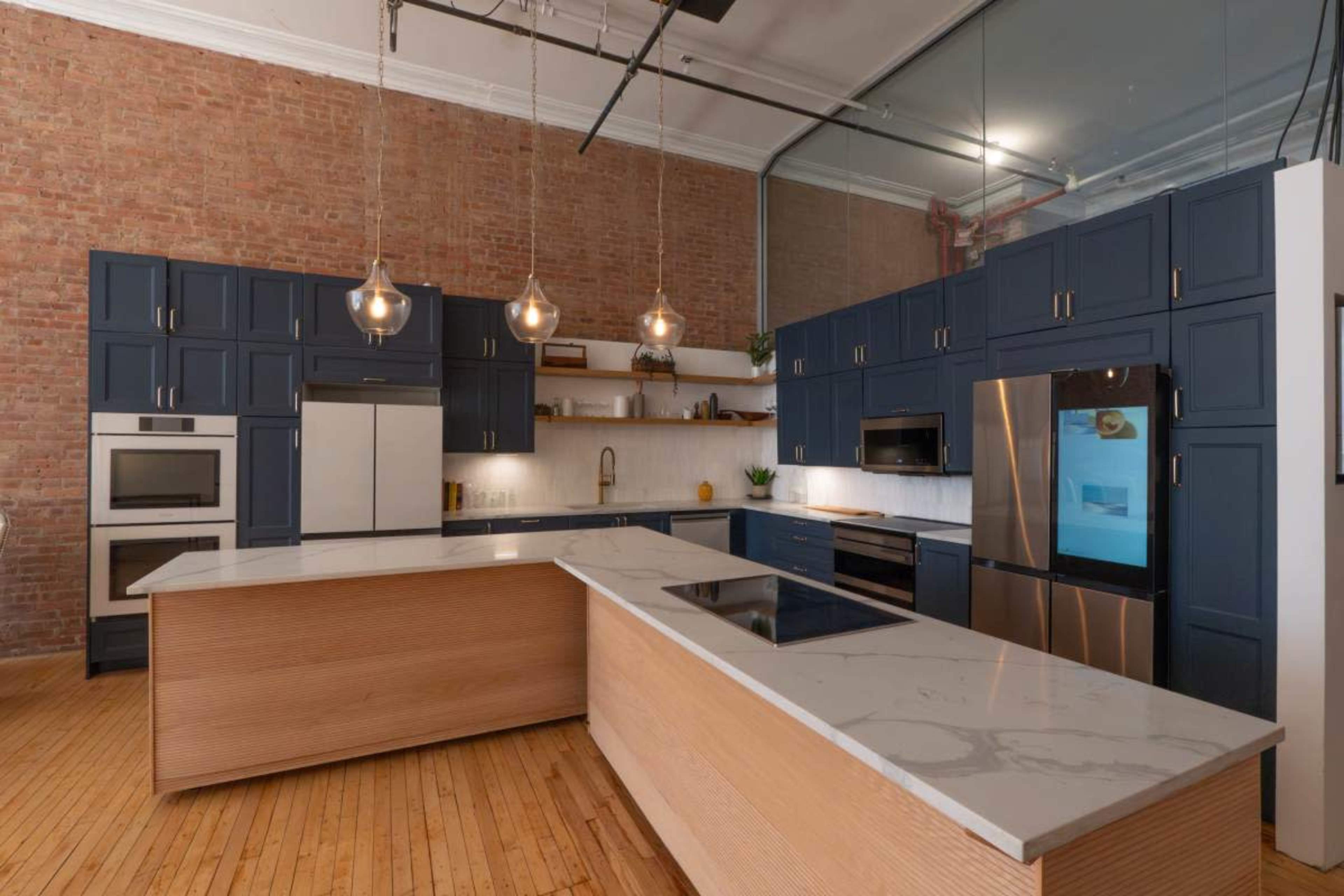 The kitchen features navy blue cabinetry, a large island with a light wood finish, and modern stainless steel appliances against a brick wall.