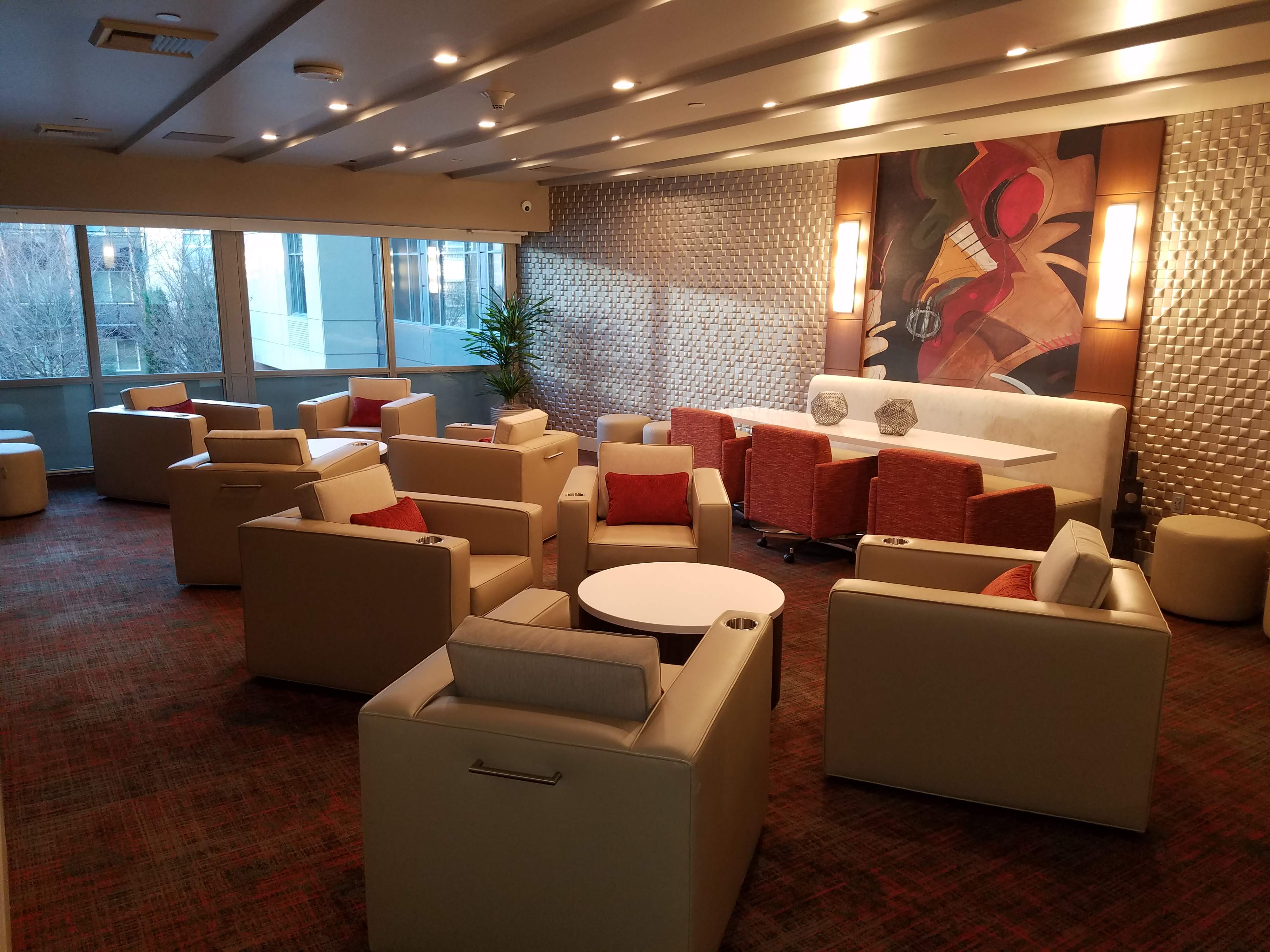A modern lounge area with several beige and orange chairs arranged around circular tables, illuminated by large windows and featuring a colorful wall art piece.