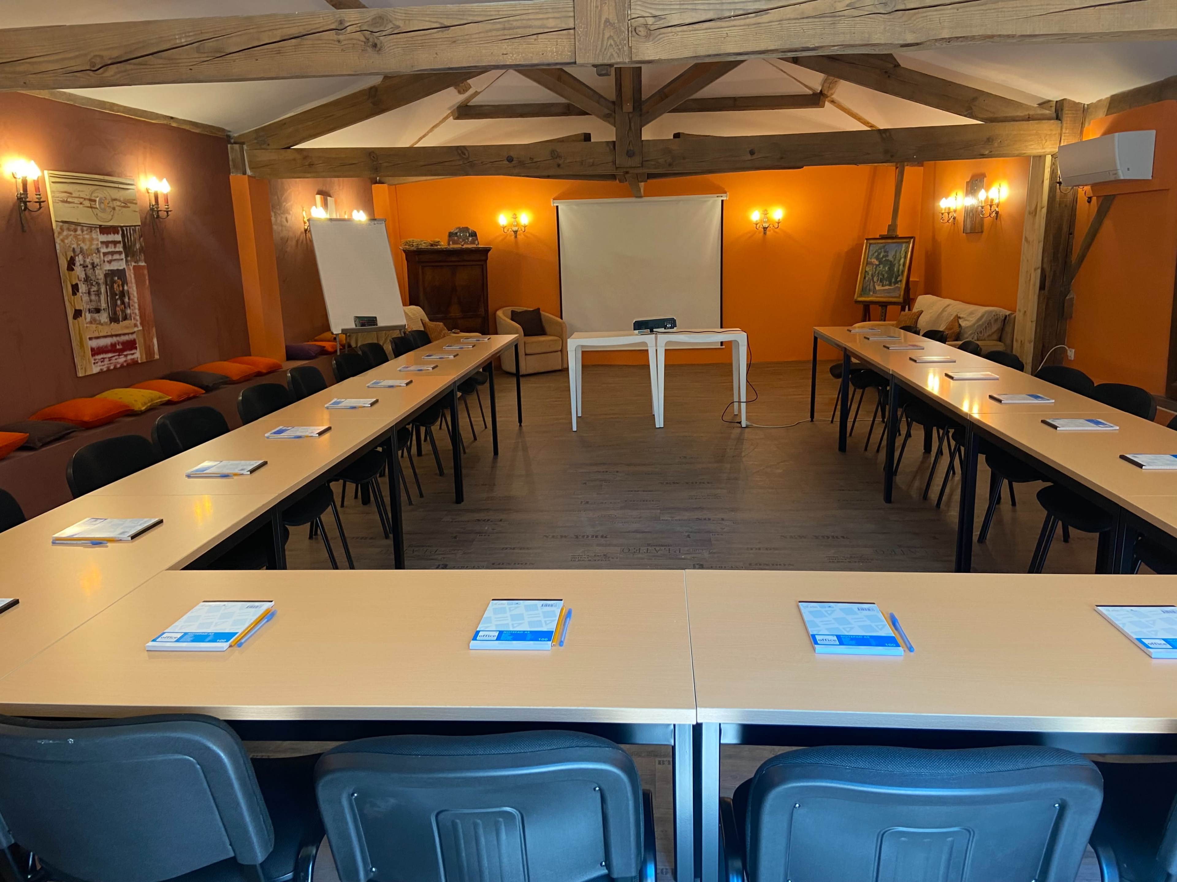 The image shows a spacious meeting room arranged with tables and chairs, featuring a projector screen at the front and flip charts on either side.