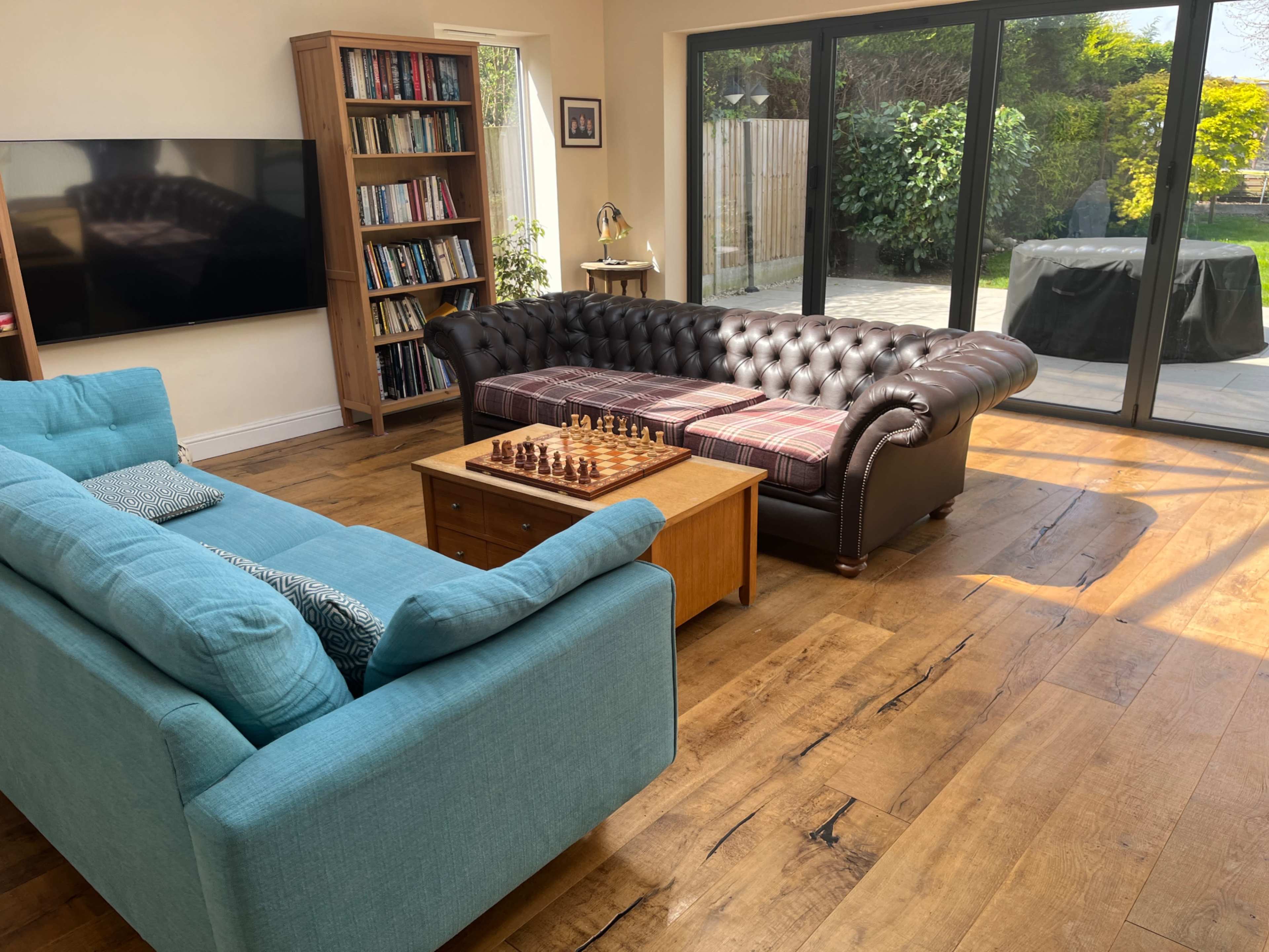 A spacious living room features a blue sofa, a black leather chesterfield couch, a wooden coffee table with a chess set, and large windows overlooking a garden.